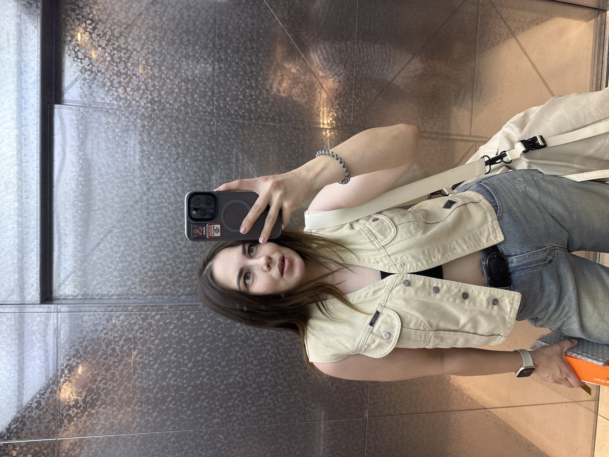 A young woman is taking a selfie in an elevator. She is wearing a beige vest over a black top, and light blue jeans. She has a white backpack slung over her shoulder. In her right hand, she is holding an orange box. She is wearing a grey coiled bracelet on her left wrist and a smartwatch on her right wrist. The elevator has metal walls. The floor is tiled. The sticker on the back of her phone says "PERM RUSSIA".