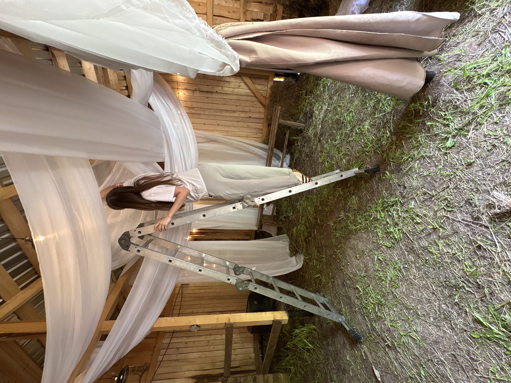 The image shows a woman standing on a silver metal ladder inside a wooden structure, possibly a barn, located outside of Sibir, Russia. She is hanging white fabric drapes from the exposed beams. The floor is covered with dry grass and dirt. A partially covered object is visible in the upper right corner. The woman has long brown hair and is wearing light-colored clothing. The lighting is a combination of natural and artificial.