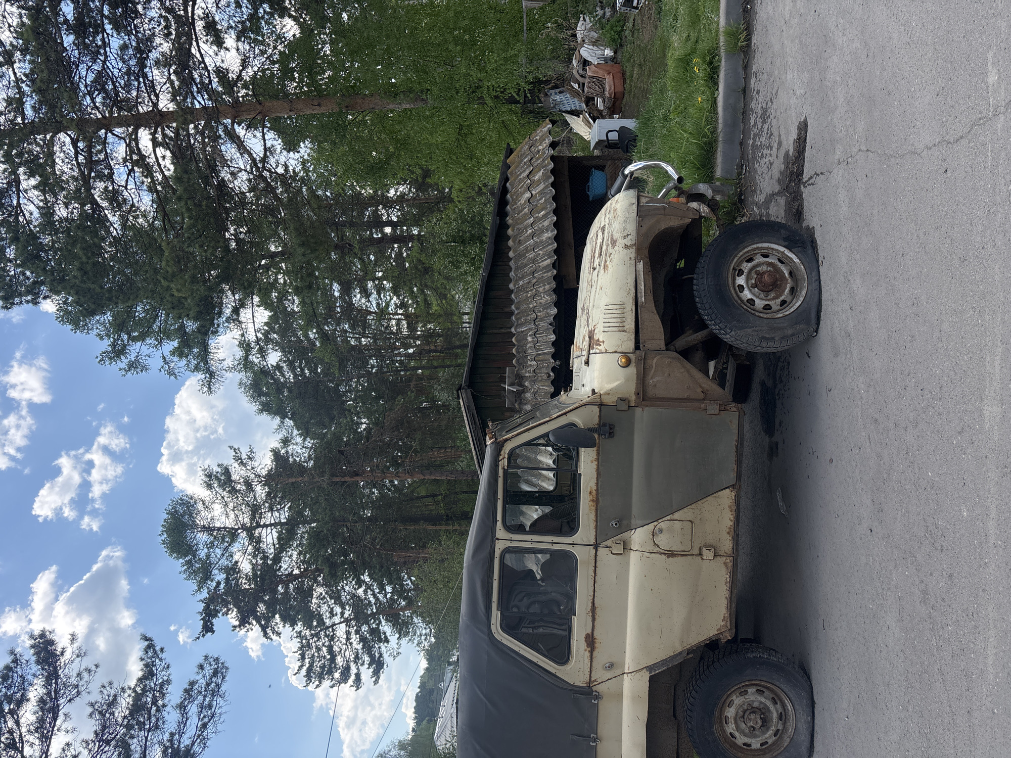 The image depicts a partially disassembled, old, off-white colored vehicle parked on the side of a paved road in Овсянка, Russia. The front passenger side tire appears flat. The vehicle is positioned near the edge of the pavement, adjacent to a grassy area and an old, weathered wooden shed. In the background, there's a collection of discarded objects and debris. A forest with tall, green trees occupies a significant portion of the background, with a blue sky and clouds visible above the trees.