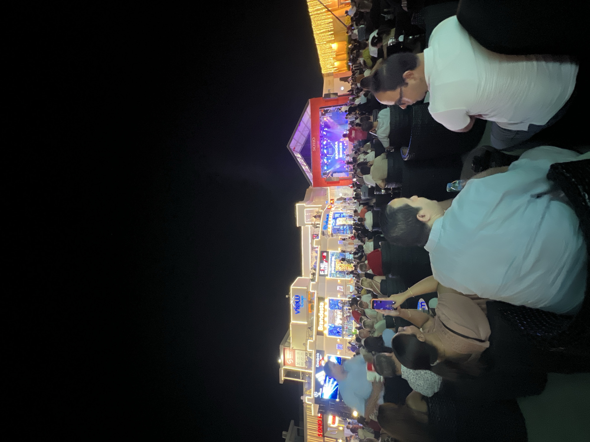 The image depicts an outdoor nighttime event at SOHO Square in Sharm El Sheikh, Egypt. A crowd of people are seated in chairs, facing a stage where a performance is taking place. The stage is lit with colorful lights and features a banner displaying "SANDRA SINGER." In the background, illuminated buildings with storefronts like "VIEW lounge," "Brandesta," and "ENE GALLERY" are visible. String lights are hung along the facade of one building. The overall atmosphere suggests a lively entertainment venue.