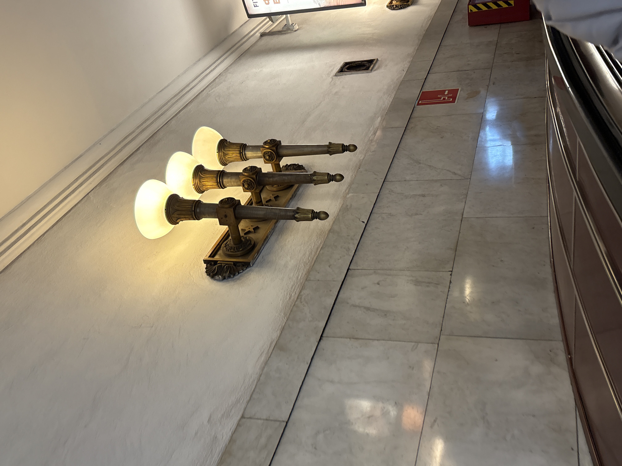 The image shows an interior scene, possibly a hallway or corridor in Saint Petersburg, Russia, characterized by its architectural details. On the left, a wall is visible, adorned with a triple-bulb sconce. The light fixtures are antique in style, made of metal, possibly bronze or brass, and have frosted glass bulbs. The wall is painted in a neutral tone, likely white or off-white, and has a slight texture.
To the right, the hallway floor is covered in marble tiles with a gray-and-white pattern. Adjacent to the floor is a dark-colored railing, indicative of an elevator or staircase. A red square with white design is on the floor. The location is likely a public building or a historic structure in Saint Petersburg.