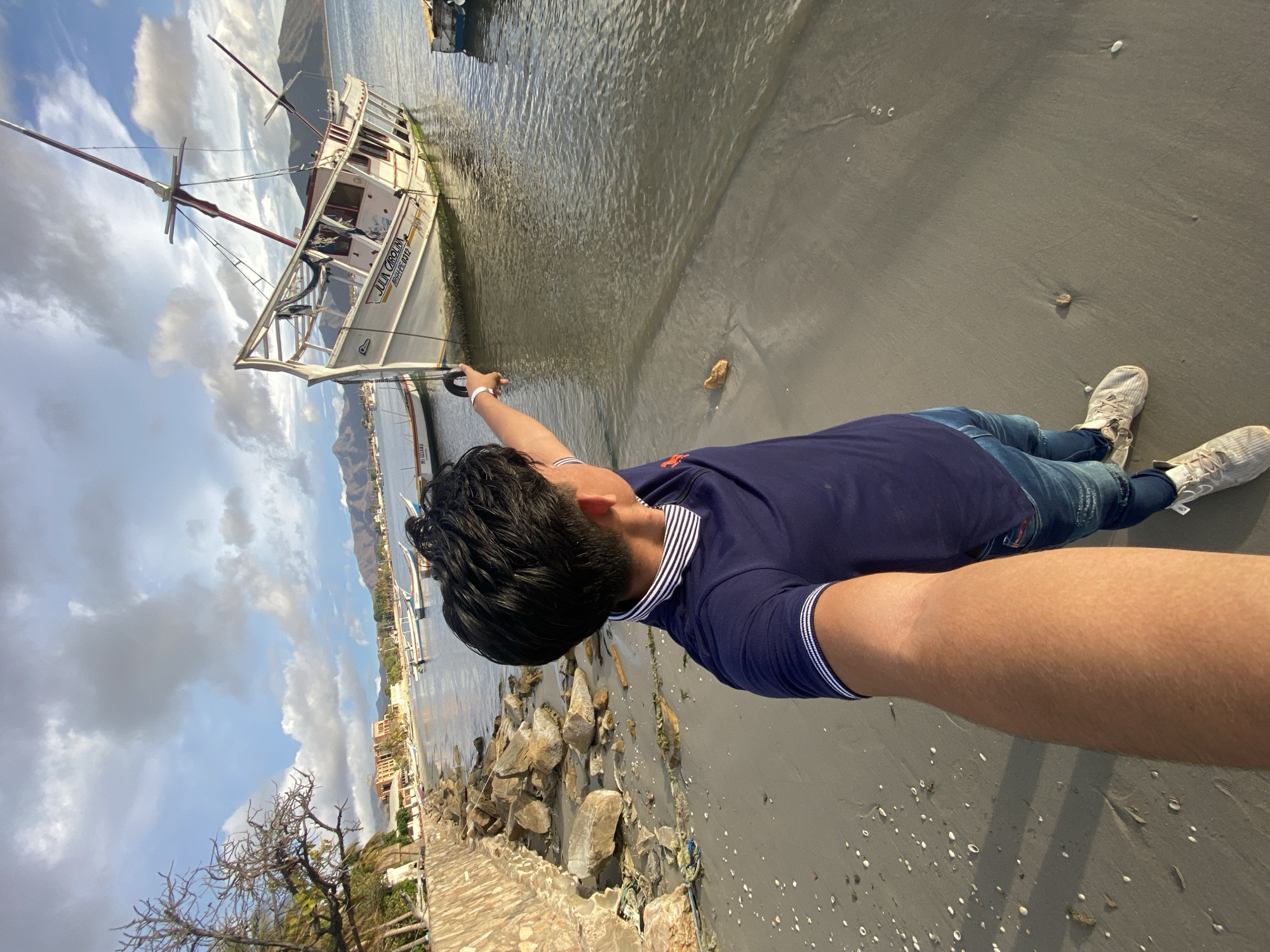 A man is taking a selfie on a beach in Juan Griego, Venezuela. He is standing near a boat on the sand, close to the water's edge. The boat is white with a red mast. The man is wearing a blue shirt, jeans, and sneakers. He's holding his arm out to take the photo. In the background, there's a town, water, cloudy sky, and mountains. The ground has rocks and sand.