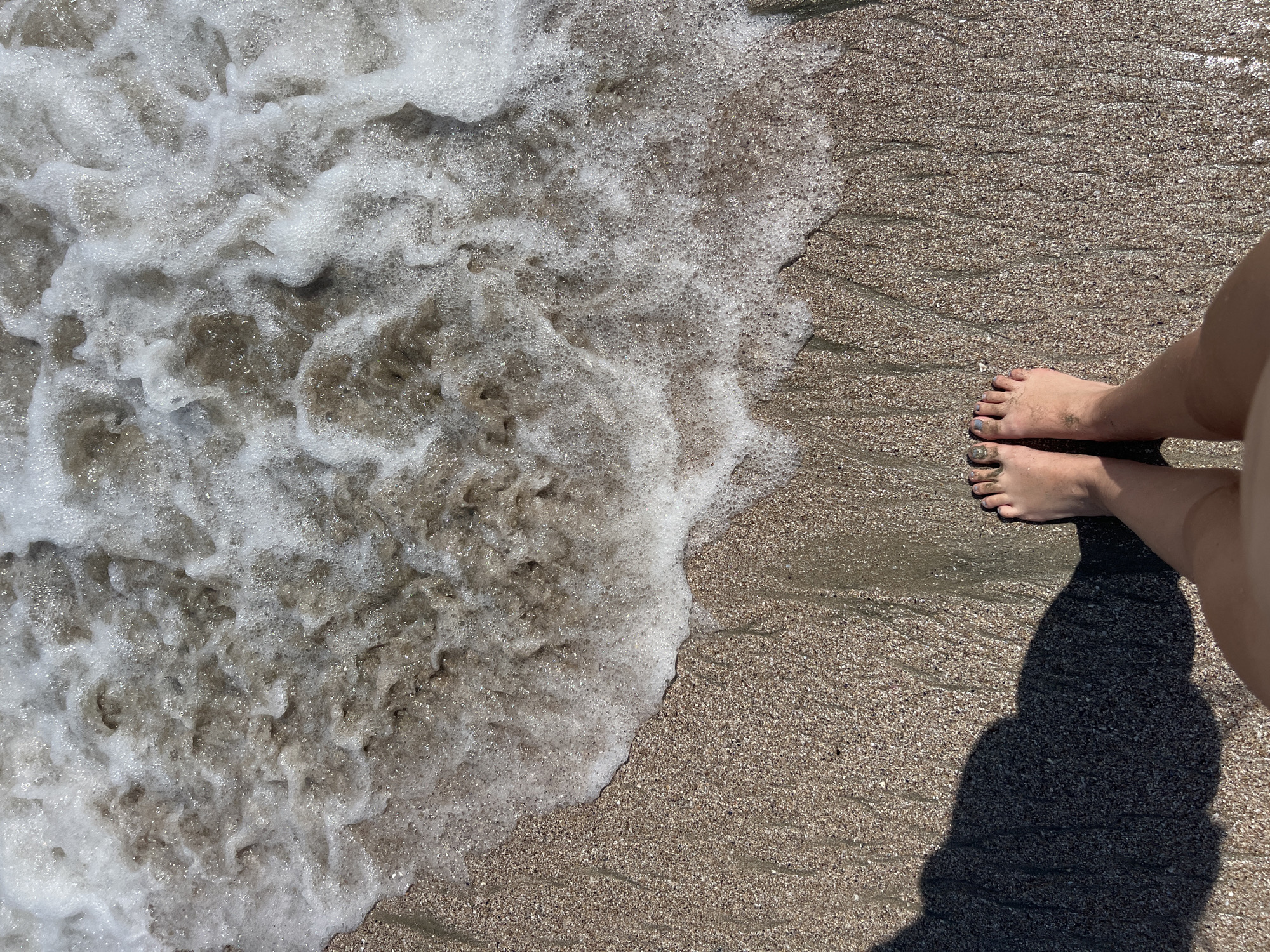 An overhead shot captures a person's bare feet standing on a sandy beach in Pomorie, Bulgaria. The feet are positioned in the lower center of the frame, with the toes of the left foot pointing slightly left and the toes of the right foot pointing slightly right. The sand is wet and compacted, with small pebbles and shells visible. A wave of foamy white water is gently lapping at the shore, partially covering the sand and reaching the toes of the person's feet. A dark shadow, cast by the person, extends from the lower left corner of the image across the sand. The overall scene evokes a sense of a relaxed moment at the seaside.