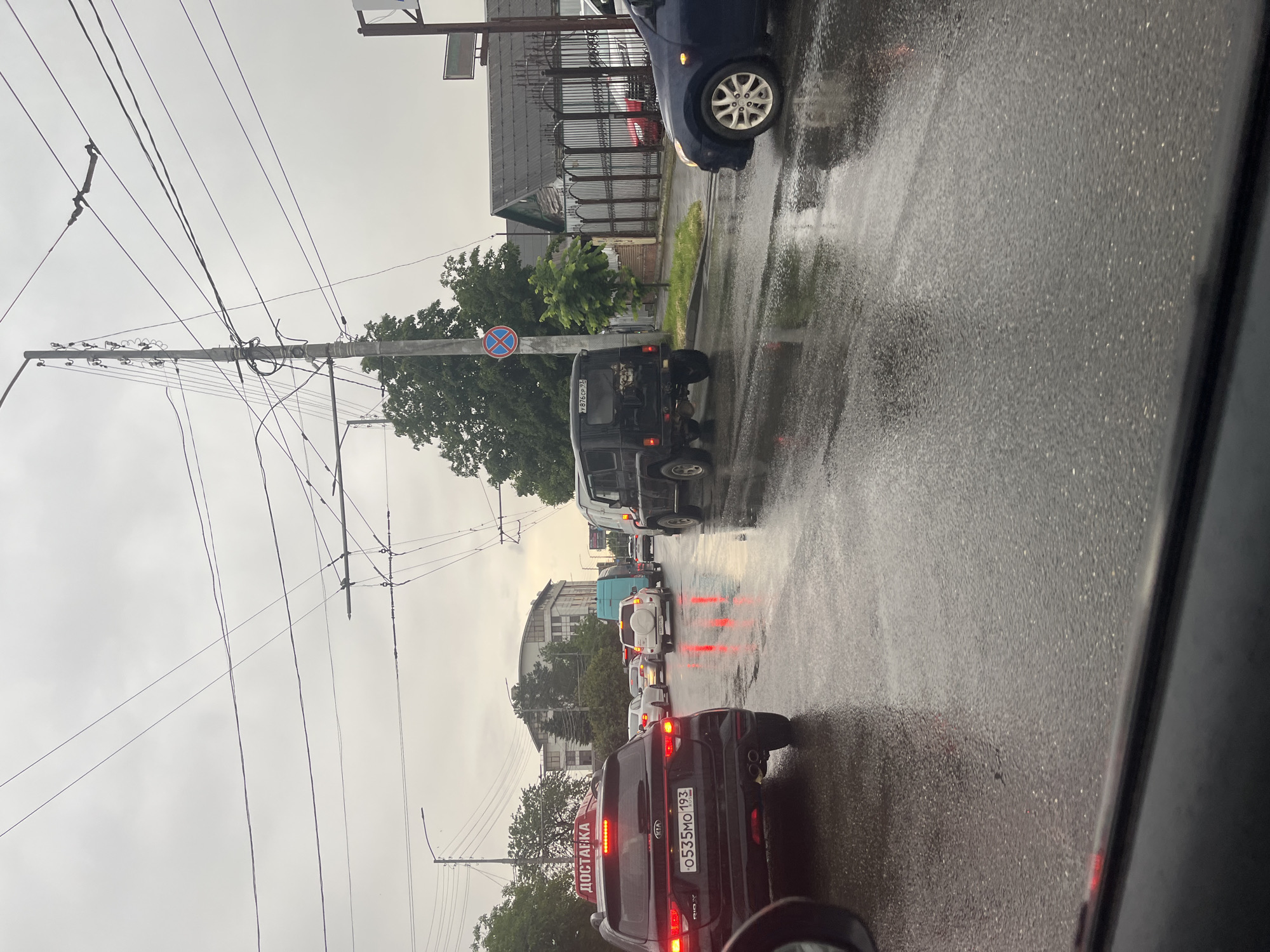 The image, taken from inside a vehicle, shows a street in Krasnodar, Russia, during rainy conditions. Several vehicles are lined up, with a dark-colored Kia SUV in the foreground displaying a license plate number 0535MO193. Visible behind it are multiple cars and a military-style truck. A road sign depicting a red circle with a line through it is visible near the truck. Overhead are power lines strung between utility poles. The environment appears to be an urban street with buildings visible in the background. The road is wet, reflecting the brake lights of the vehicles.