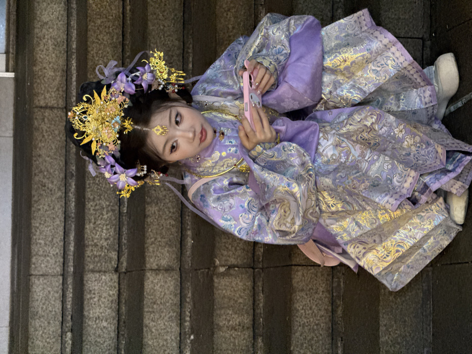 A young East Asian female is observed squatting on a series of grey, rough-hewn stone steps in Yuzhong District, China. She is attired in an elaborate traditional Chinese costume, predominantly lavender with silver and gold brocade patterns, featuring a high collar and voluminous sleeves. Her head is adorned with an ornate golden headpiece, which includes a central crown-like structure, additional golden ornaments, and purple floral embellishments. A small golden ornament rests on her forehead, and a gold-colored necklace is visible. She holds a light pink smartphone horizontally with both hands, her thumbs positioned on the screen, indicating active use. Her gaze is directed slightly to the viewer's left. The environment is dimly lit.