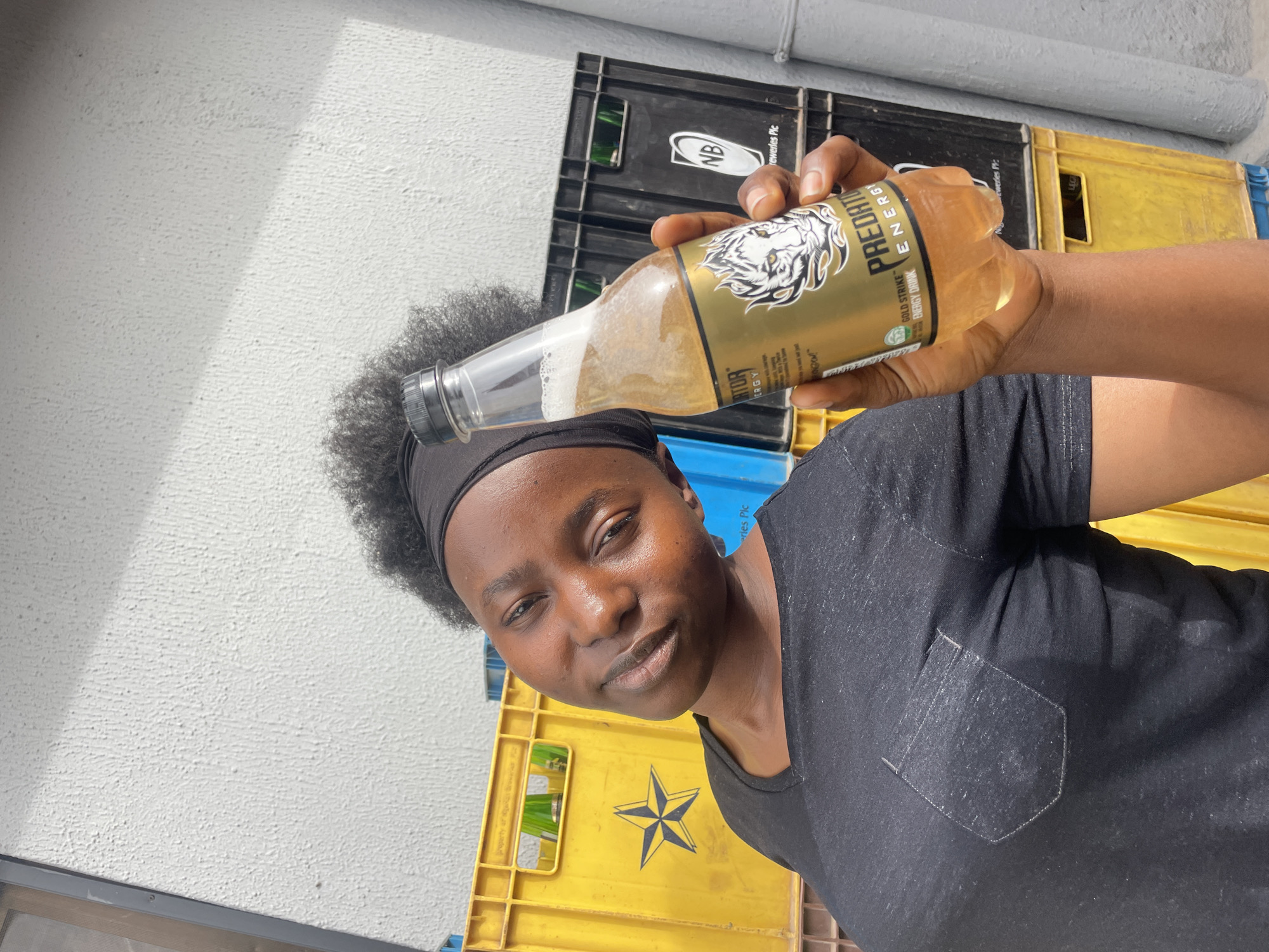 A young person, appearing to be a dark-skinned individual with short, curly black hair and a black headband, is depicted in a tilted, close-up shot. They are looking directly at the camera with a slight smile and relaxed expression. The person is wearing a black t-shirt with a small pocket on the chest.
They are holding up a clear glass bottle containing a golden-brown, frothy liquid, likely an energy drink. The bottle has a prominent gold and black label featuring a white tiger head logo. Visible text on the label includes "PREDATOR ENERGY" and "GOLD STRIKE ENERGY DRINK."
The background features a textured white wall, strongly lit suggesting daytime and sunny weather, with some shadows cast. Behind the person and the bottle, there are stacks of industrial-style crates. Some are black with "NB" and "Breweries Plc" visible, indicating a connection to Nigerian Breweries. Other crates are yellow, one of which displays a black star logo. Given the context of Jos, Nigeria, these details are consistent with local industry. The overall scene appears to be outdoors or in a semi-enclosed industrial area.