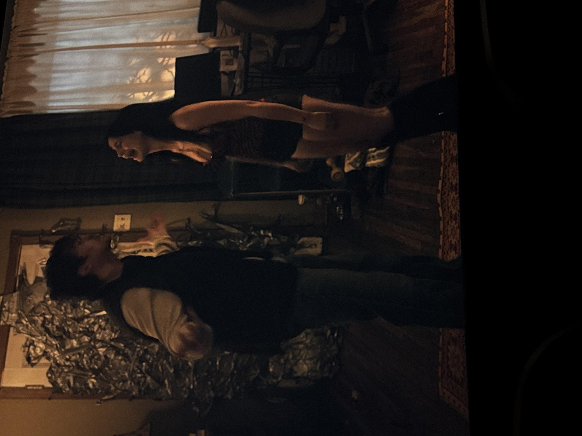 A man in a white long-sleeve shirt and black vest stands with arms raised in front of a window covered in foil. A woman stands facing him, turned slightly to the right, and is looking away from him. Both individuals are indoors. The scene appears to be a room with a patterned rug visible in the foreground. The location is unknown and is not possible to discern from the image.