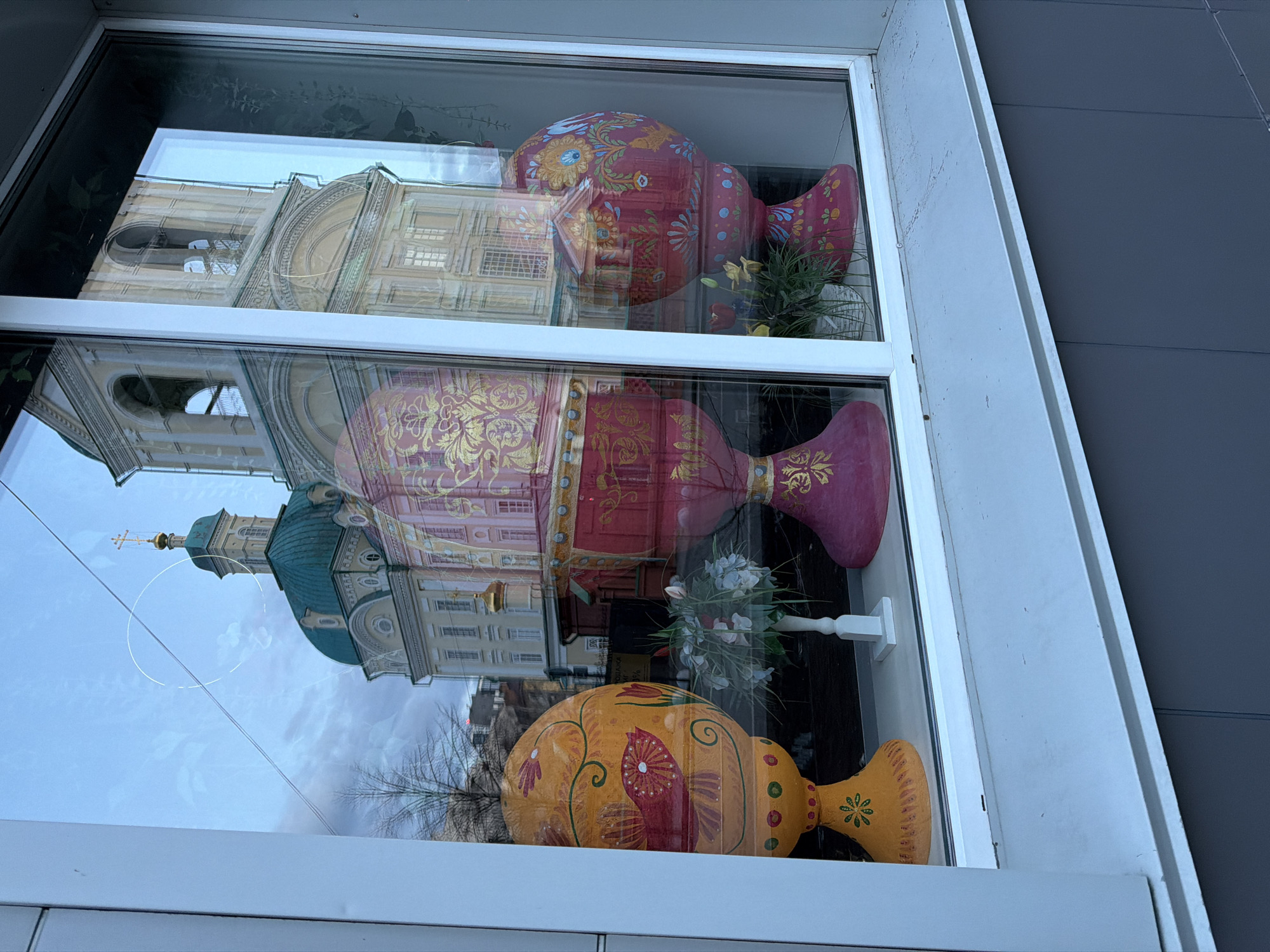 The image shows a window display in Krasnoyarsk, Russia, featuring three large, decorative Easter eggs. The eggs are stacked vertically, with a yellow egg at the bottom, a red egg in the middle, and another red egg at the top. Each egg is uniquely painted with floral and geometric patterns. The window reflects the Church of the Intercession of the Mother of God in Krasnoyarsk, revealing its architectural style with light-colored walls, arched windows, and a blue and green dome. Between the eggs are small bouquets of artificial flowers in a white vase. The window frame is white, and a gray wall is visible on the right side of the image.