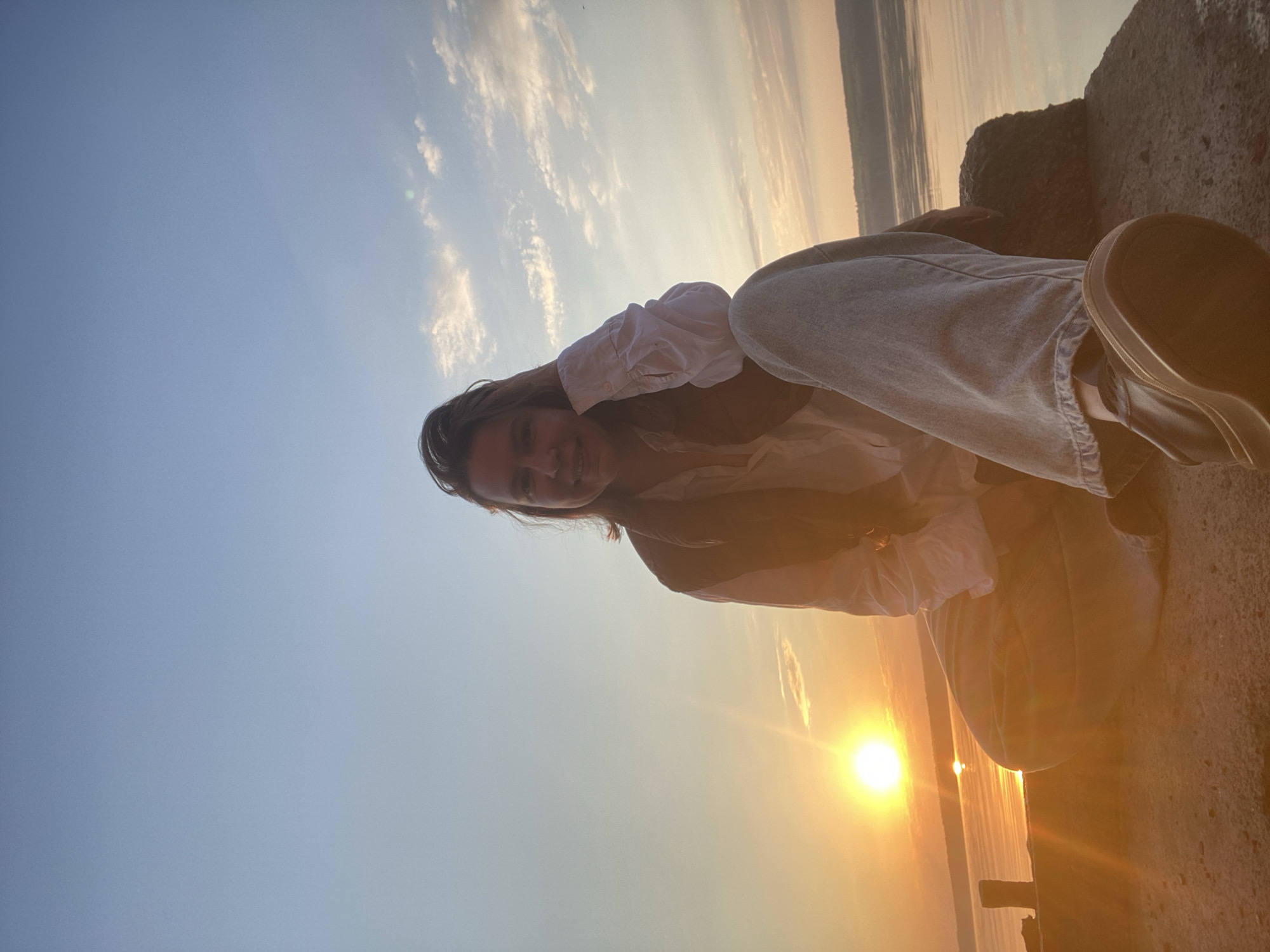 A young woman is photographed at sunset in what appears to be the city of Chaikovsky, Russia, given the horizon line visible in the photo. She is sitting on a stone structure, possibly a breakwater or pier. She is wearing a white collared shirt, a vest, and jeans. The sun is setting over water. The sky is blue near the top of the image, fading to orange near the horizon.