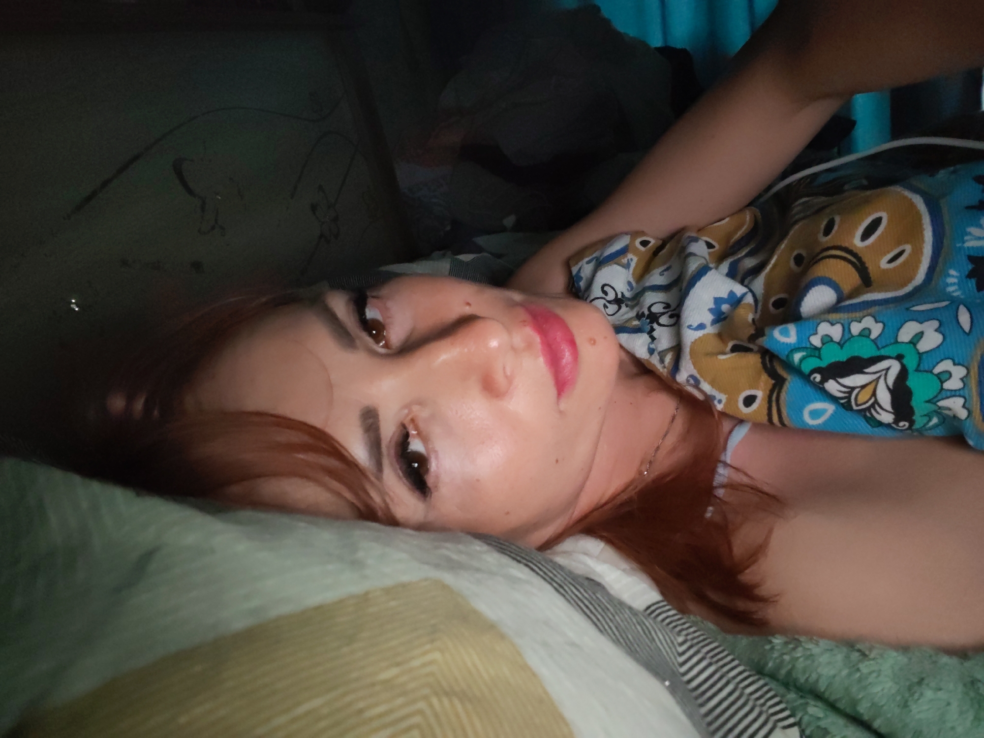 The image shows a woman lying on a bed with her head tilted to the side. She has red hair and is wearing makeup, including red lipstick. Her eyes are open and looking towards the camera. She is wearing a patterned top. The background is dimly lit and appears to be a bedroom. There are no discernible landmarks or features that would indicate the specific location within Chaikovsky, Russia. The overall impression is a candid, personal photograph.