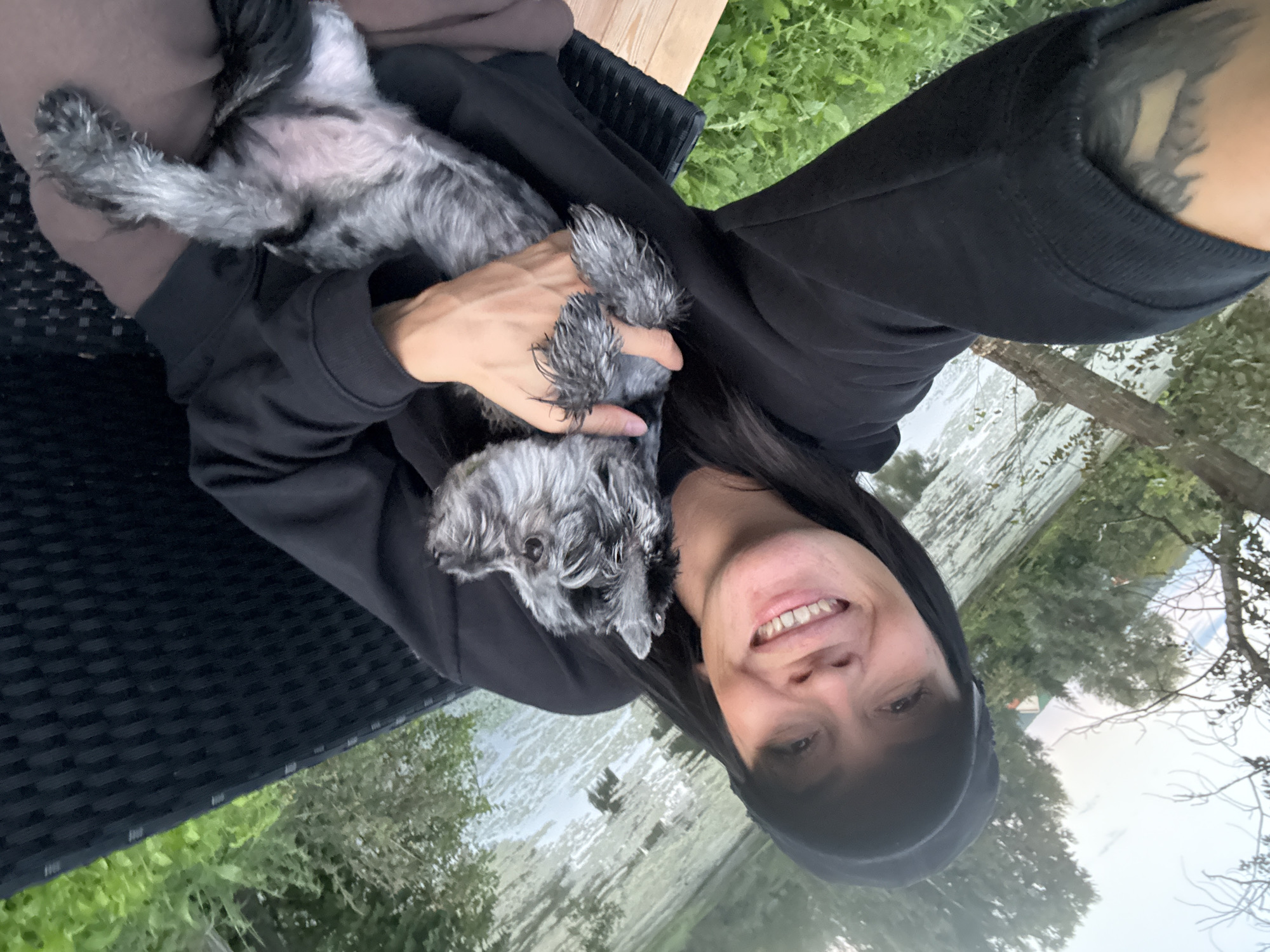 A person wearing a black hooded sweatshirt holds a small gray schnauzer dog in their arms. The person is smiling and looking at the camera, while the dog appears to be resting. They are seated on a dark wicker chair with a wooden frame. In the background, there is a body of water, trees, and green grass, suggesting an outdoor, possibly park-like setting. The scene is likely located in Chaikovsky, Russia, given the context provided.
