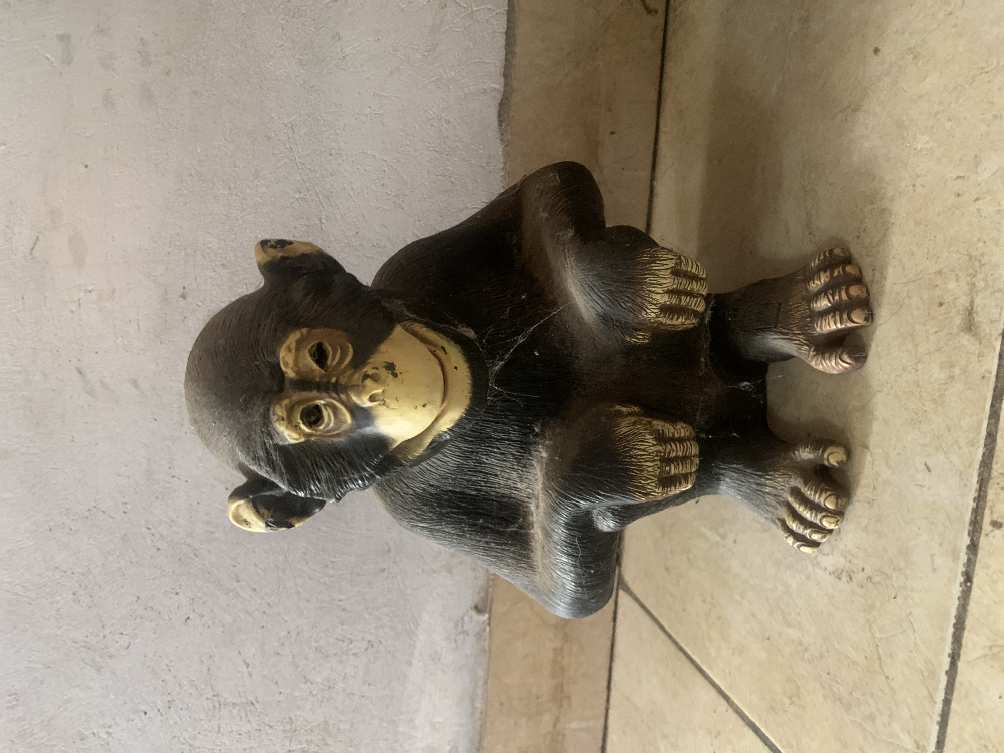 A slightly-worn monkey figurine is seen sitting on a tiled floor next to a painted wall. The monkey is dark grey or black with yellow accents around the face, hands, and feet. There are signs of wear and small cobwebs on the figurine. The tiled floor has a light beige color, and the wall is light gray. The location is not discernable as the environment is minimal. There is no indication of this scene being in Santa Ana, Venezuela.