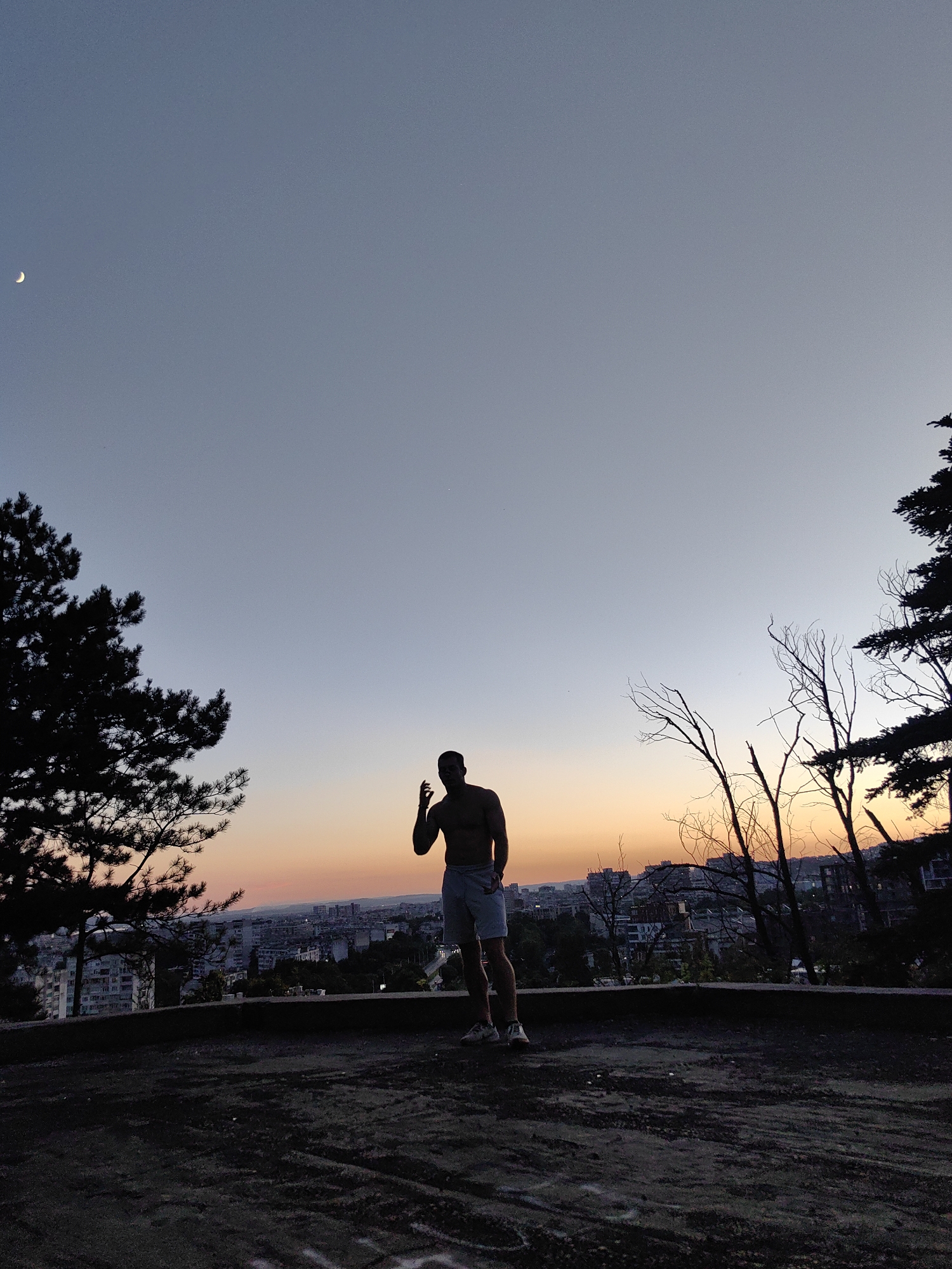 A silhouette of a shirtless man stands in the foreground, facing a city skyline at dusk. The man is wearing shorts and athletic shoes. He has his right hand raised, with his index finger extended. Behind him, the sky transitions from a pale blue at the top to a vibrant orange and pink near the horizon, indicating a sunset. A crescent moon is visible in the upper left portion of the sky. The city below is characterized by buildings of various sizes and heights. Trees, some with sparse foliage and others dense, frame the scene on the left and right sides. The ground in the foreground appears to be a dark, textured surface, possibly pavement or dirt. The image suggests a viewpoint from an elevated position overlooking the city. This viewpoint is consistent with a location in the city of Varna, Bulgaria, such as a hill or a higher vantage point offering panoramic views.