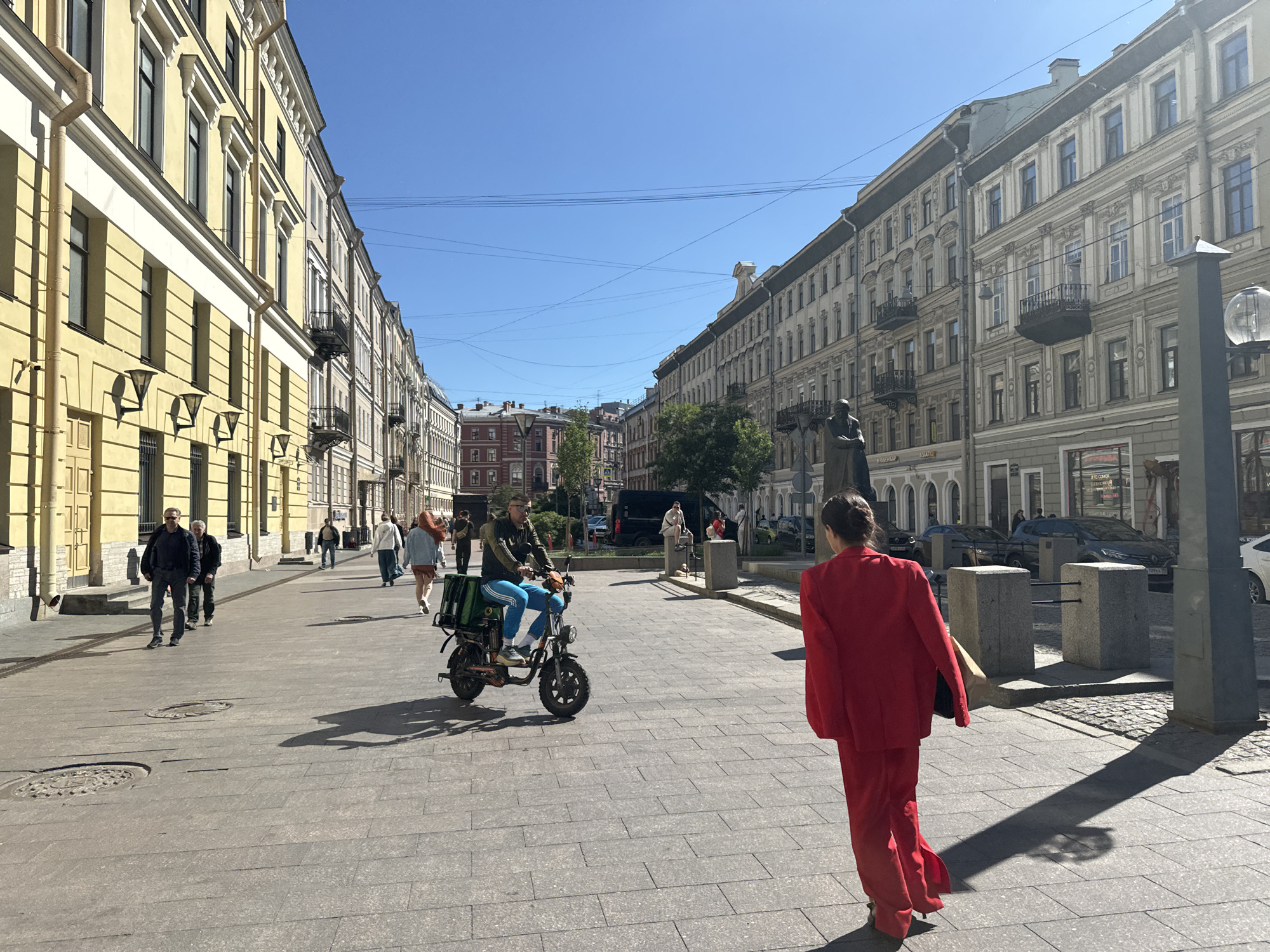 This is a medium-shot, eye-level, outdoor photograph taken on a sunny day on a city street in Saint Petersburg, Russia. The scene depicts a wide, paved pedestrian area lined with historic buildings. In the foreground, a woman in a bright red suit walks away from the camera, casting a long shadow on the pavement. To the left, several people are walking, including a man in a dark jacket and blue striped pants riding a scooter with green bags attached to the back. Further down the street, more pedestrians are visible, as well as a few parked cars. The buildings on either side of the street are multi-story, with ornate architectural details and balconies. The sky is clear and blue. The overall impression is of a bustling urban environment on a pleasant day.