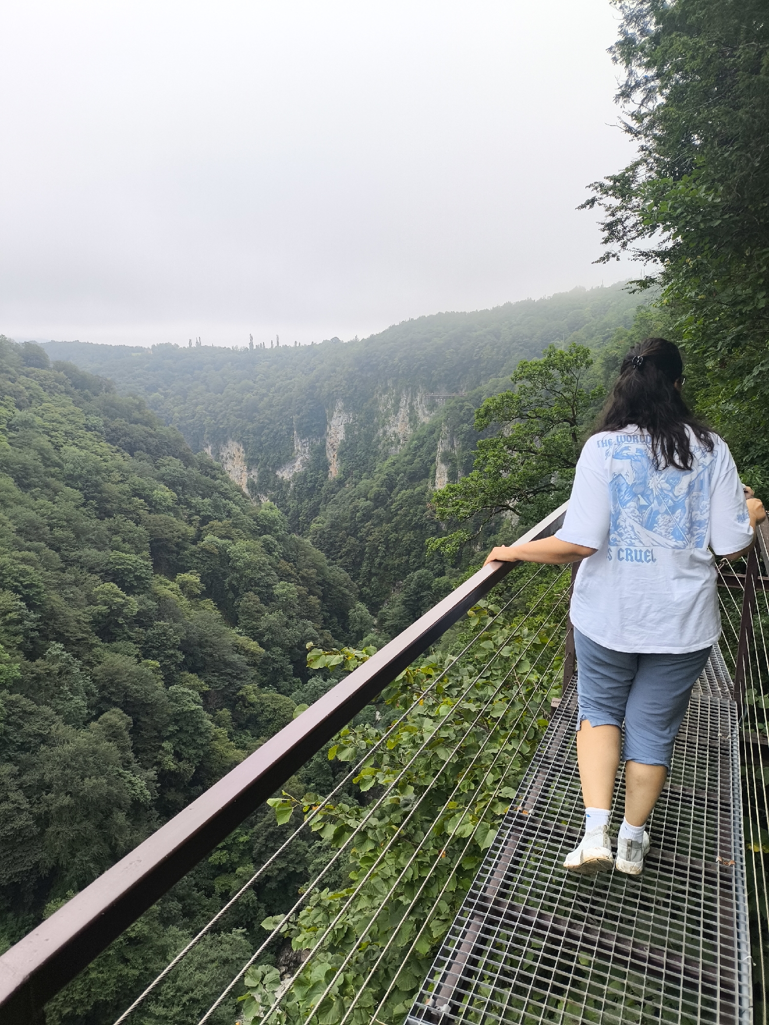 A woman with dark hair tied back with a clip walks along a metal grate walkway on a bridge. She is facing away from the camera, wearing a white t-shirt with a blue graphic and text that reads "THE WORLD IS CRUEL", and light blue capri-length pants. She is wearing white athletic shoes and socks. Her right hand rests on the metal railing of the bridge, and her left hand is slightly behind her, also near the railing. The walkway is bordered by a dark brown railing on the left and a cable railing on the right, with dense green foliage growing on either side.
The bridge overlooks a vast, deep canyon filled with lush green trees. The canyon walls are steep and rocky, with some exposed rock faces visible. The sky is overcast and a light mist hangs in the distance, obscuring the far side of the canyon and the horizon. The overall environment is a natural, mountainous landscape. The location is identified as the Martvili Canyon in Satsiskvilo, Georgia, a popular tourist destination known for its dramatic geological formations and natural beauty.