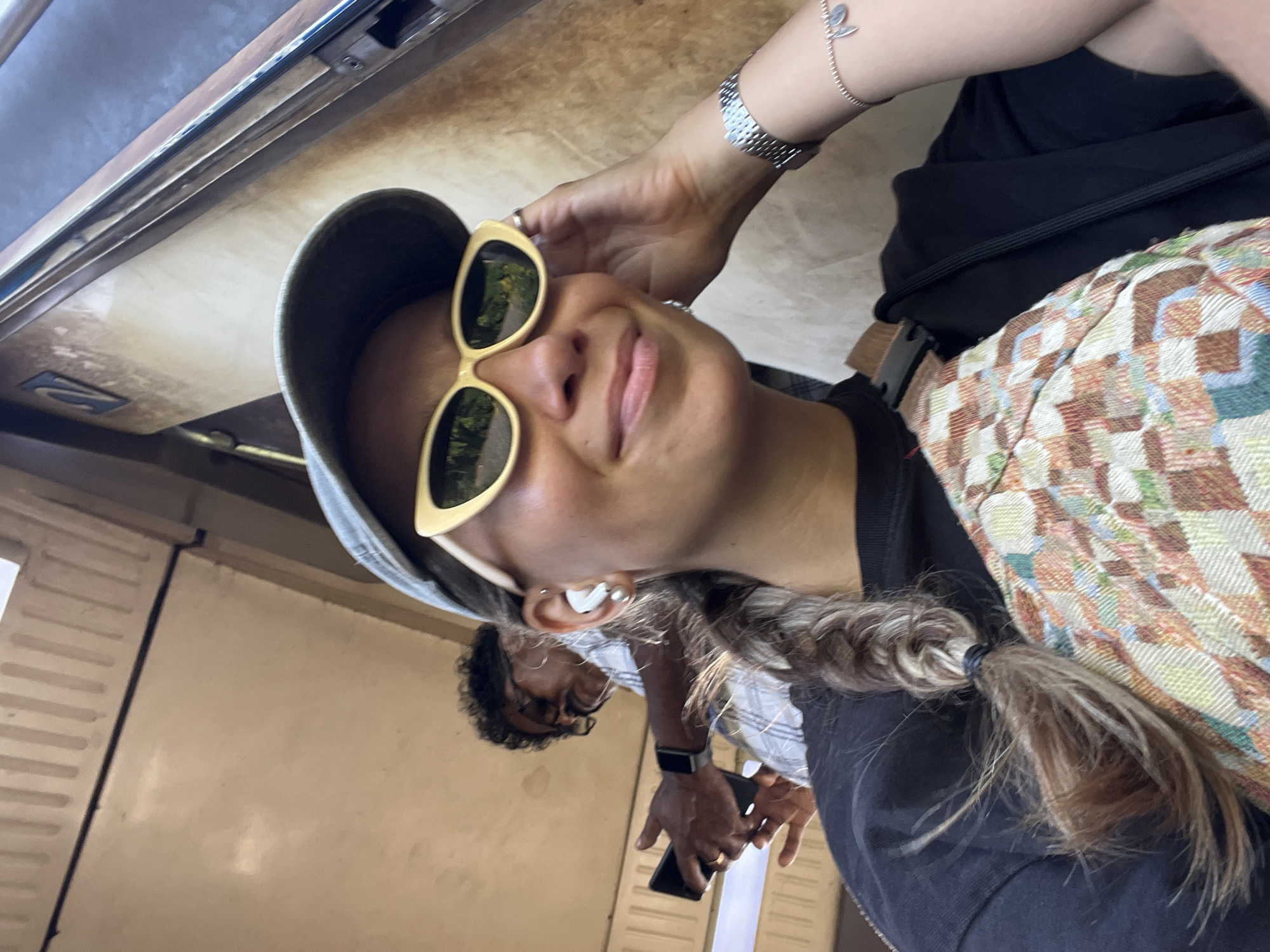 The image is a selfie taken by a woman wearing a grey baseball cap, yellow sunglasses, and a black t-shirt. She has a braid of hair extending over her shoulder and is wearing white wireless earbuds. A silver watch is visible on her right wrist, along with a bracelet with a charm. Behind her, a man wearing glasses and a checkered shirt is holding a black smartphone. The background appears to be the interior of a train carriage, with worn beige walls and vertical ribbed panels. A sign with the number "2" is visible on the wall behind the man. The overall lighting suggests it is daytime. This scene is consistent with being inside a train carriage of the Sri Lanka Railways, likely operating on a route that passes through Kalutara, a city known for its coastal train line.