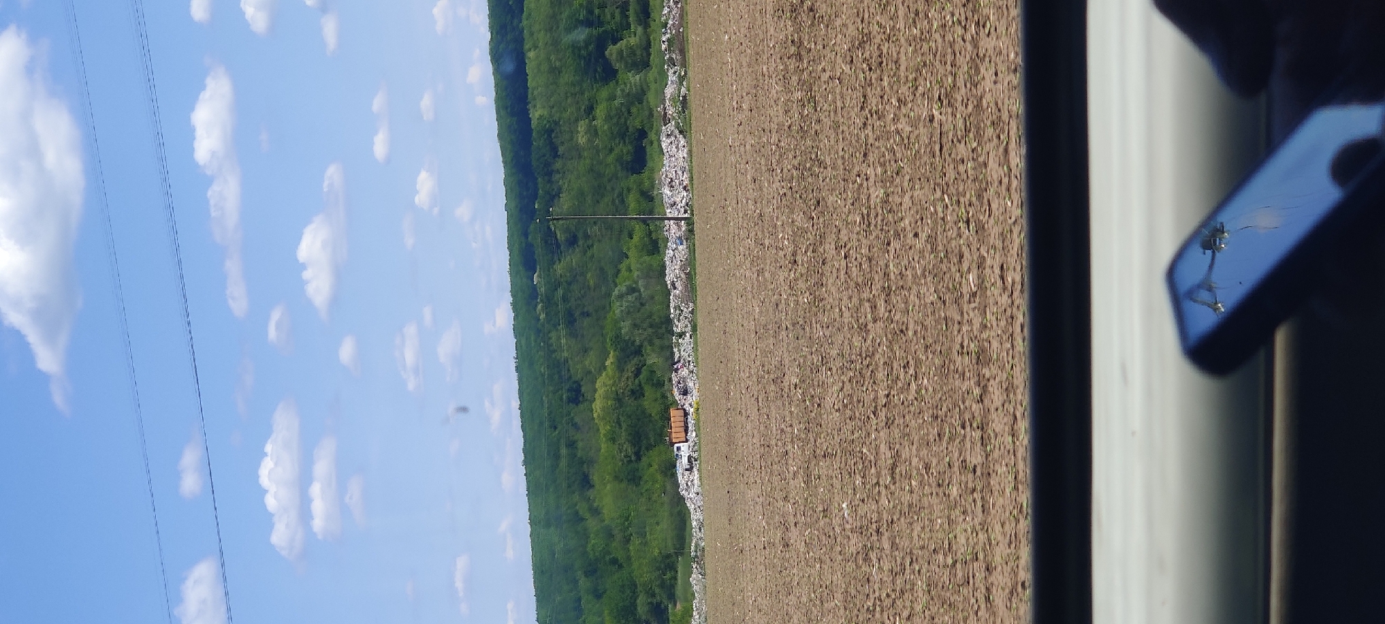 The image captures a wide outdoor landscape under a bright, clear blue sky adorned with numerous fluffy white clouds. Two parallel power lines traverse the upper portion of the sky.
In the middle ground, a long, dense band of green forest stretches across the horizon, likely covering a low hill or ridge. Immediately in front of this forest line, and extending into the mid-ground, is a very large, elongated pile of white and light-colored material, strongly suggesting a landfill or a significant accumulation of waste. A single utility pole stands upright within or directly in front of this large waste pile. Towards the right side of this waste area, a white truck with a brown or wooden rectangular container on its bed is visible.
The foreground is dominated by a vast, tilled agricultural field of brown earth, exhibiting faint linear patterns from cultivation. Small green sprouts are sparsely visible across the field.
On the far right edge of the frame, a dark, vertical structure, likely a window frame or part of a vehicle's interior, partially obstructs the view. Reflected in this surface is a blurry image of a hand holding a dark-colored smartphone, indicating the photograph was taken from inside, likely a moving vehicle, looking out.
The scene depicts a sunny day with good visibility, presenting a rural or exurban environment where cultivated land, natural forest, and a substantial waste disposal site coexist. No discernible text is visible.