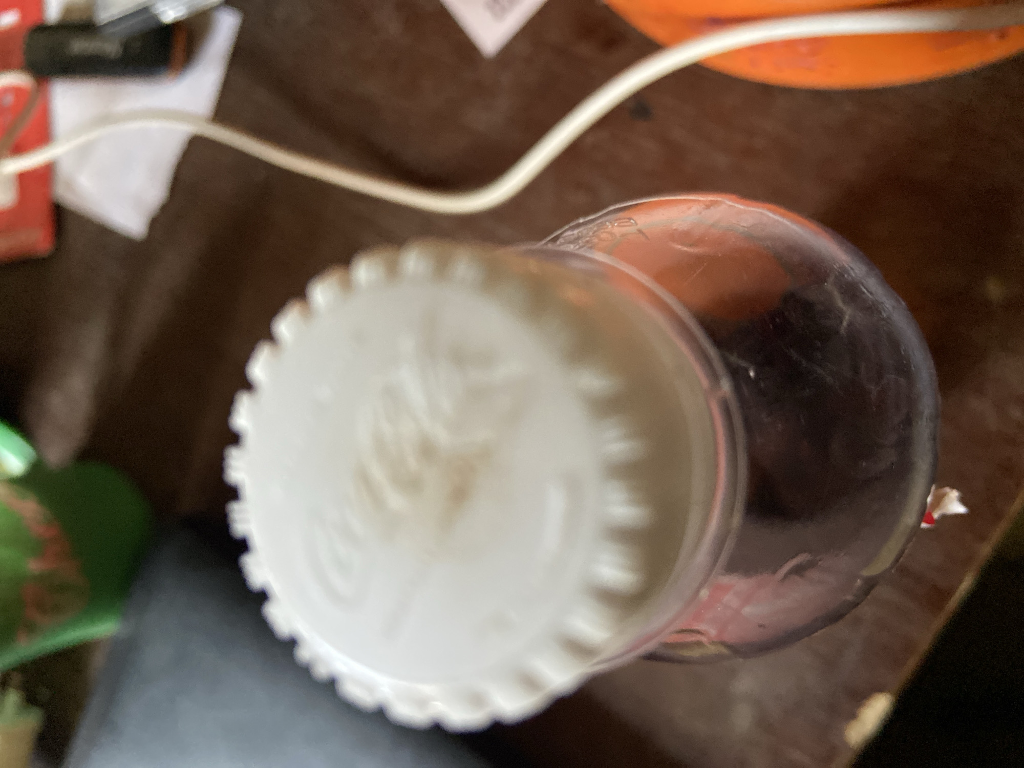 The scene is an indoor setting in Mararaba, Nigeria, displaying a close-up, high-angle view of objects on a surface. Dominating the center is a transparent plastic bottle filled with a dark, opaque liquid, topped with a white, ribbed screw cap. The cap's surface features subtle, indistinct embossed markings and minor light brown discoloration. A small piece of red and white material is visible at the very bottom of the bottle. To the left of the bottle, a dark, matte surface extends, with a blurred green container featuring red markings in the background. To the right of the bottle, a white cable, thick and curved, descends from the upper right. Adjacent to the cable, a blurred, rounded orange object is present. Further in the background to the right, a dark brown surface is visible, along with a piece of white paper or tissue, a dark cylindrical object, and a partially obscured red rectangular item, all out of focus. No individuals are depicted, and no active interactions are occurring.