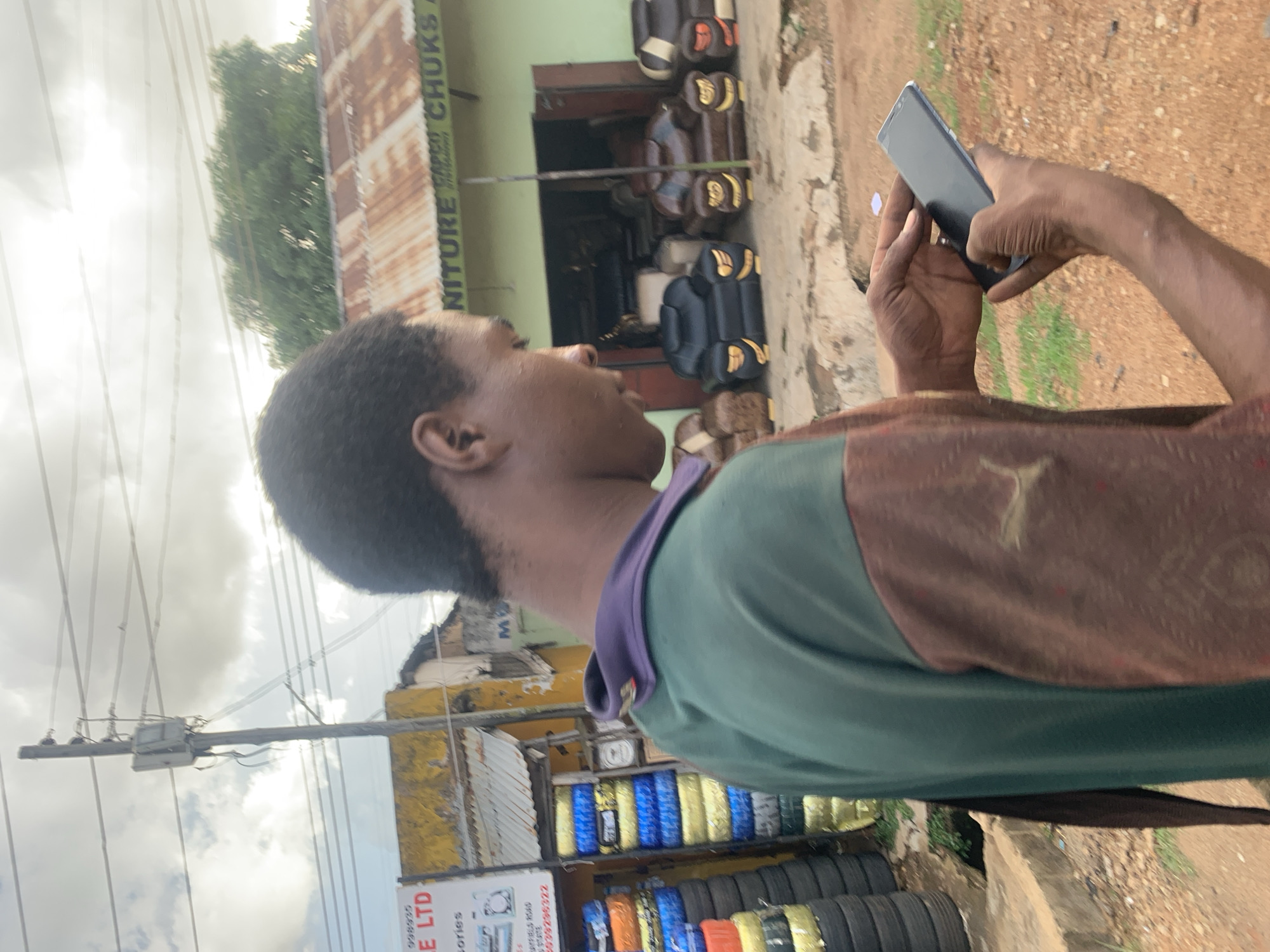 This media file shows a person holding a smartphone. The person is a dark-skinned individual with short, dark hair, wearing a green shirt with a brown yoke. They are standing outdoors, with a building behind them that appears to be a shop. The building has a corrugated metal roof and a sign that reads "NITURE CHUKS". Inside the shop, several chairs and other items are visible. In the foreground, there is a display of tires, some of which are brightly colored. The ground is a mix of dirt and pavement, with some sparse vegetation. The sky is overcast with clouds. The image seems to have been taken during the daytime. The person is looking at their phone, suggesting they might be using it for communication, entertainment, or perhaps to take a photo or video. The overall scene depicts a street-side vendor or shop in what appears to be a bustling urban or semi-urban environment. The presence of tires and furniture suggests a commercial area. The image is oriented vertically, with the person's head at the top and their feet towards the bottom.