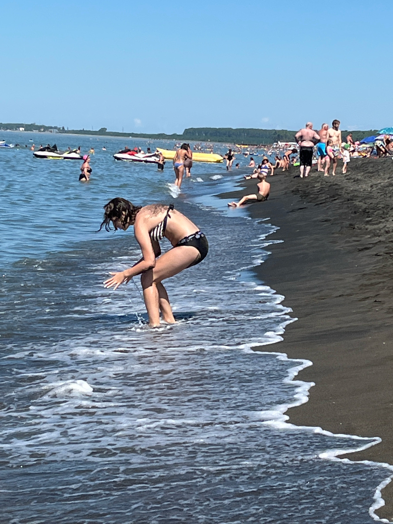 A beach scene in Ureki, Georgia features a young woman in a black and white striped bikini bending down with her hands in the shallow water. She has dark, wet hair. The dark sand beach extends to the right, with numerous people visible in the background. Some are standing, others are sitting or lying down. Several people are in the water, including some on jet skis and a yellow inflatable tube. The sky is clear and blue. The water has small waves breaking on the shore.