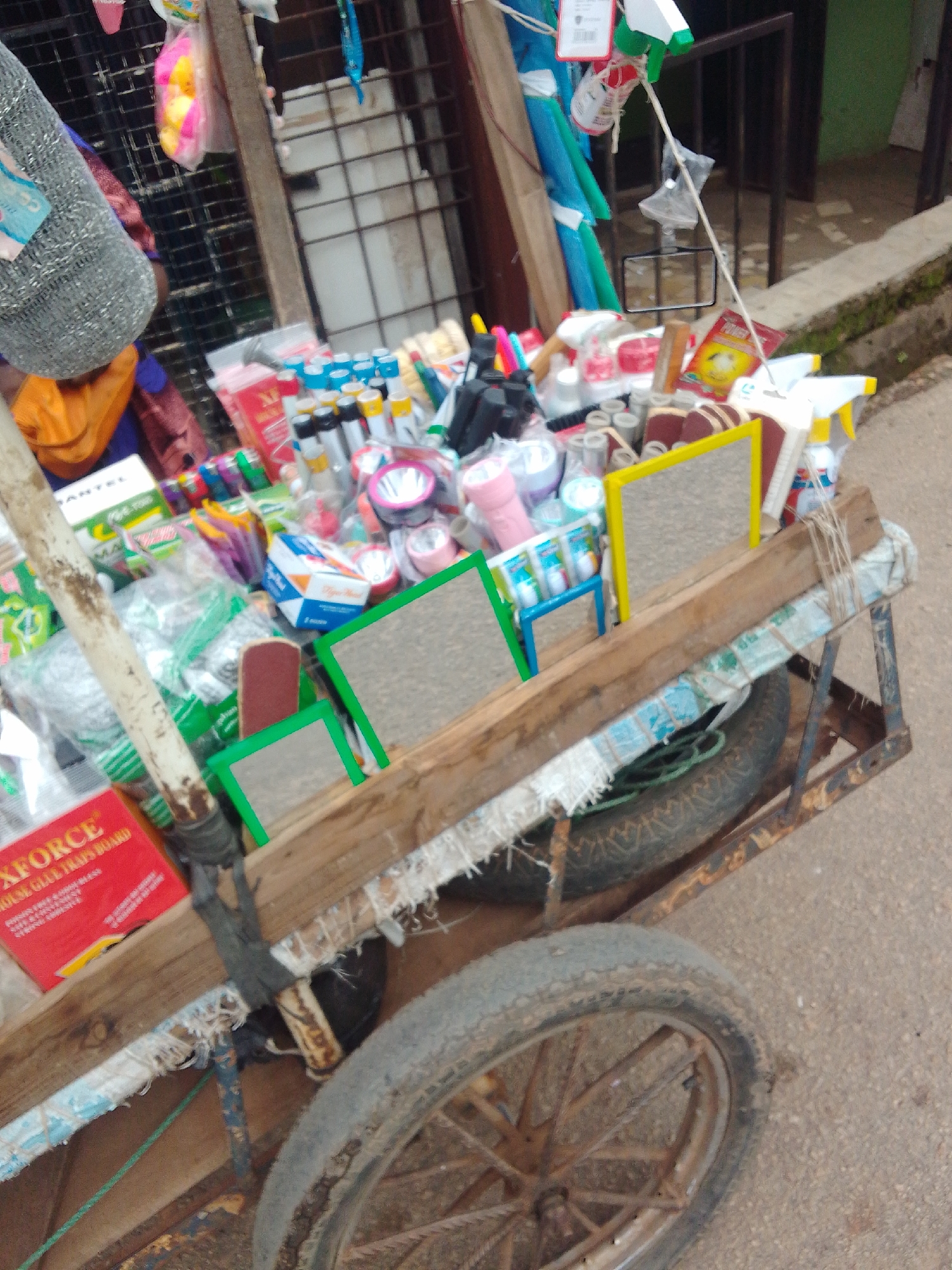 This image captures a heavily loaded, weathered pushcart, likely belonging to a street vendor, situated on a paved surface in the city of Jos, Nigeria. The cart's structure consists of a rusted metal frame supporting a platform made of worn wooden planks. Two spoked metal wheels, heavily corroded and fitted with worn pneumatic tires, are visible, with the front wheel prominent in the foreground. A robust, stained wooden pole extends vertically from the front of the cart, secured with dark material, serving as a handle or support.
The cart is densely packed with an assortment of small consumer goods. Visible items include: a red box labeled "XFORCE MOUSE GLUE TRAPS BOARD"; various small handheld mirrors with green and yellow frames; numerous small bottles and containers with multi-colored caps (blue, white, yellow, black); several flashlights, including pink and white models; plastic spray bottles with green and yellow nozzles; packaged items that resemble rolls of sandpaper; assorted plastic tools, brushes, and colorful clips; and various other packaged products in clear plastic bags, including green bags and a white box partially labeled "MANTEL". A red package labeled "POWER" is also discernible within the merchandise.
The immediate environment consists of a light brown, paved ground surface, suggesting asphalt or concrete. The background on the left reveals a metal grid or gate, while the right side features a building structure with a dark metal gate, a concrete ledge showing moss growth, and a section of a green-painted wall. Additional miscellaneous items, such as a grey woven bag, a blue elongated object, and a pink fluffy item, are visible hanging in the distant background. No individuals are present within the visible frame, suggesting the cart is stationary, possibly awaiting customers or in transit.
