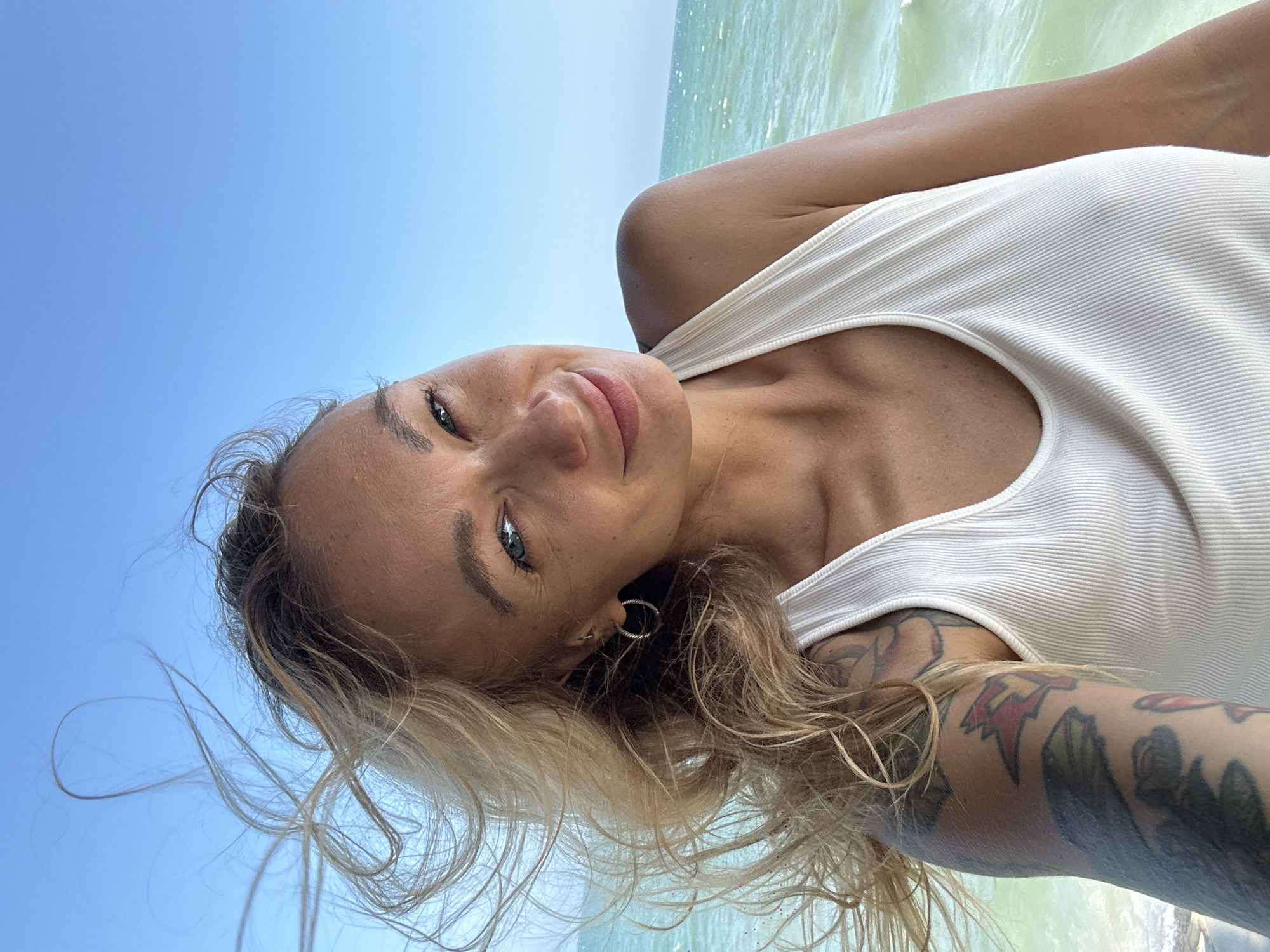 A female subject is captured in a head-and-shoulders selfie photograph, smiling directly at the camera. Her long, blonde hair is actively blowing to the viewer's right, indicative of strong wind. She has light-colored irises and faint freckles on her face, which is slightly tilted. She is wearing a ribbed, off-white halter-neck top. Visible on her left arm are extensive, multi-colored tattoos. The background consists of a clear, bright blue sky in the upper left and a light green, somewhat choppy body of water with small waves in the middle and right, suggesting a coastal or beachfront environment. The scene is illuminated by natural daylight. This scene is depicted in Phu Thuy Ward, Vietnam.
