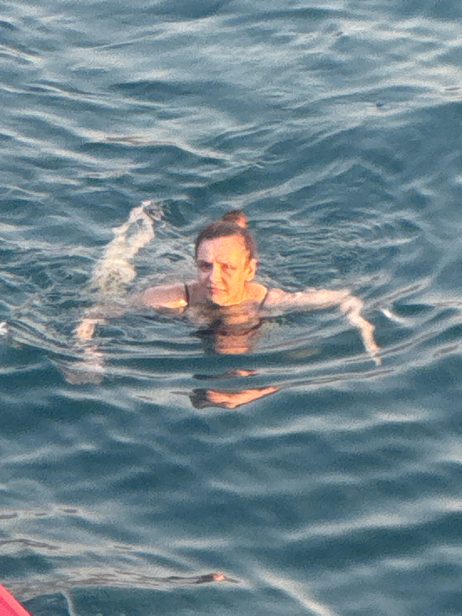 A woman is swimming in the sea in Kamenovo, Montenegro. She is in the center of the frame, facing the camera, with her head and shoulders visible above the water. Her hair is pulled back into a bun, and she is wearing a dark-colored swimsuit. The water is a deep blue-green color and is slightly choppy, with waves creating ripples and reflections. The lighting suggests it is daytime, possibly late afternoon or early morning, as there are warm tones on the woman's face. Part of a red object, possibly a boat or float, is visible in the lower-left corner of the image. The background is entirely composed of the sea.
