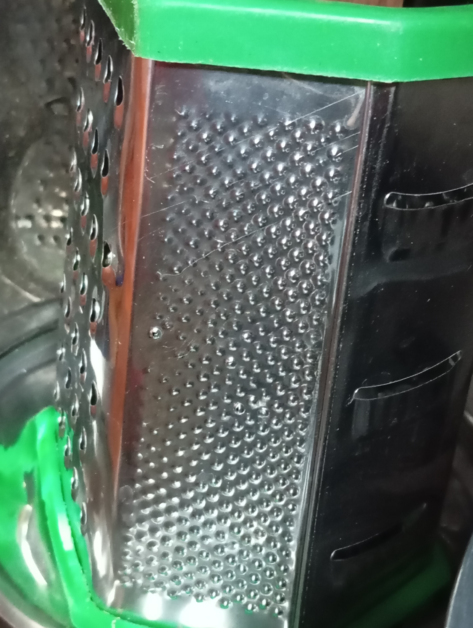 A close-up, eye-level shot shows a stainless steel box grater with a green plastic top. The grater has four sides, two of which are visible. The side on the left has small, oval-shaped perforations, while the side on the right has larger, curved slots. The surface of the grater is reflective and shows some scratches and smudges. A portion of what appears to be a green plastic handle is visible on the left side of the frame. The background is dark and out of focus, with hints of metallic surfaces and green plastic. The grater is positioned vertically. The image is taken indoors, likely in a kitchen or food preparation area. There are no people or any other objects clearly identifiable in the scene.