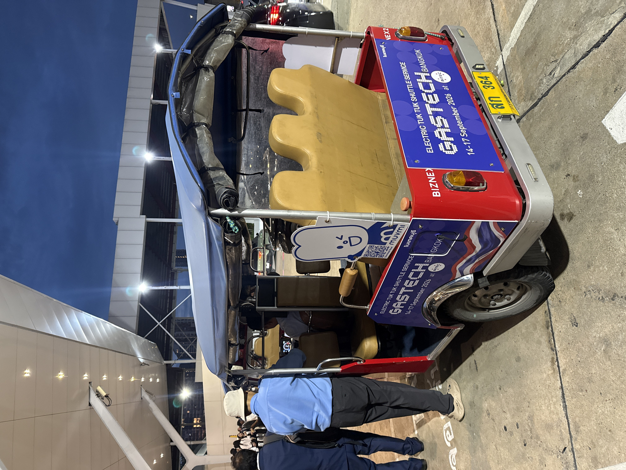 The image shows a red and blue electric tuk-tuk shuttle service vehicle parked on a concrete surface, likely an airport or event venue, in Bangkok, Thailand. The tuk-tuk features promotional signage for "GASTECH" and "BIZNEX," indicating an event scheduled for September 14-17, 2026, at BITEC, Bangkok. A man in a blue shirt and a hat is standing next to the driver's side door, and another man in a suit is walking away from the tuk-tuk. In the background, a modern building structure with white panels and artificial lighting is visible against a darkening twilight sky. The overall scene appears to be an outdoor area at dusk or evening.