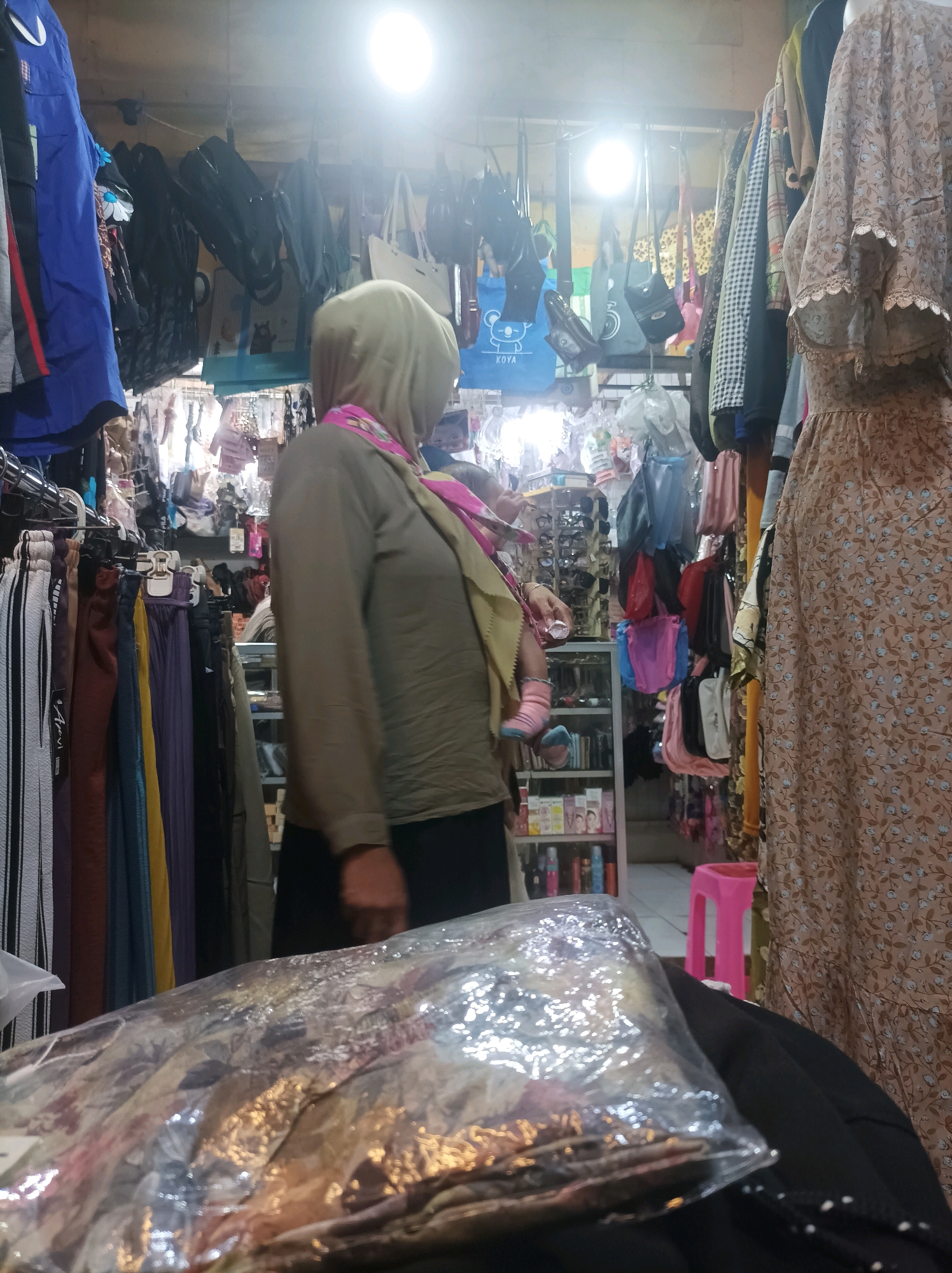 A woman wearing a hijab and a pink scarf is in a market in Sumuragung, Indonesia. She is holding a baby, also wearing a hijab. The market is full of clothes and bags hanging from the ceiling and on racks. There are also shelves filled with various items. The lighting is bright and uneven. The woman and baby are the main subjects of the image, and their interaction is the focus. The market environment provides context for their presence.