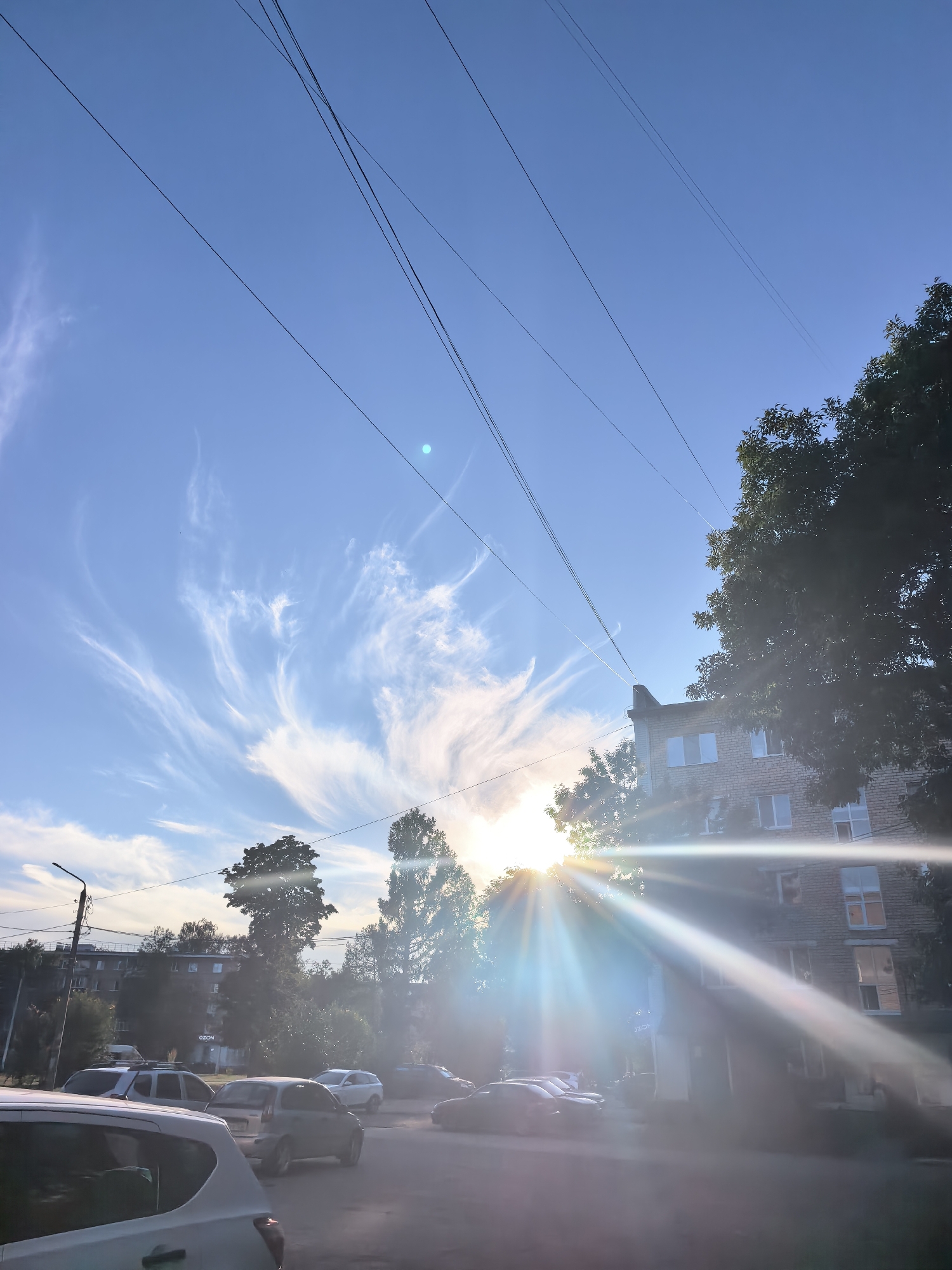The image depicts a street scene in Sterlitamak, Russia, bathed in the warm light of a setting or rising sun. The sky is a vibrant blue, streaked with wispy white clouds that catch the sunlight, creating a dramatic effect. Power lines crisscross the sky, adding a utilitarian element to the natural beauty.
In the foreground, several cars are parked along the street, suggesting a residential or commercial area. A white car is prominently visible on the left, its rear end angled towards the viewer. Other vehicles, including a silver hatchback and a darker SUV, are also present.
Behind the cars, a multi-story brick building stands, its windows reflecting the bright sky. Trees with lush green foliage are scattered throughout the scene, some towering over the buildings, while others are smaller and interspersed with the architecture. The sun, partially obscured by trees and the building, casts long, bright rays that create a lens flare effect, illuminating the scene with a golden glow. A street lamp is visible on the left side of the image. The overall impression is one of a peaceful urban environment experiencing a beautiful natural phenomenon.