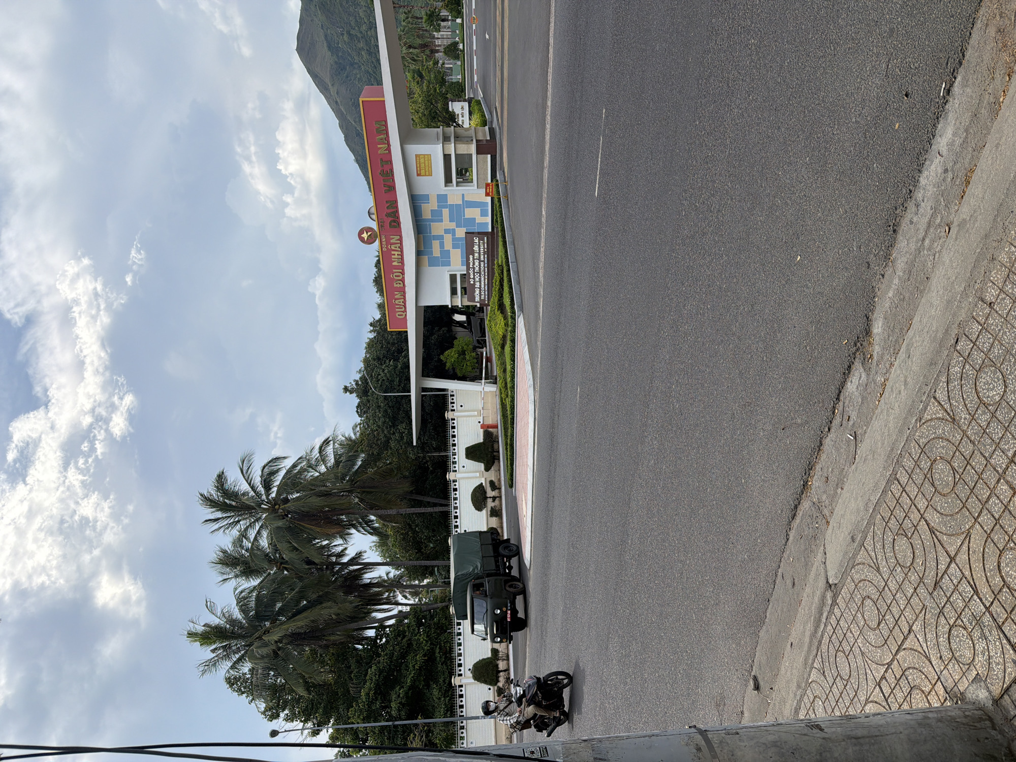 A military entrance to the People's Army of Vietnam is shown. The entrance features a large red sign with yellow lettering that reads "QUÂN ĐỘI NHÂN DÂN VIỆT NAM". Below this, a smaller sign is visible that states "BỘ QUỐC PHÒNG TRƯỜNG ĐẠI HỌC THÔNG TIN LIÊN LẠC TELECOMMUNICATION UNIVERSITY". Palm trees line the entrance. A green military truck is seen entering the premises. In the foreground, a person on a motorcycle is traveling on the road. The sky is cloudy, and a mountain can be seen in the background. The scene is located in Bac Nha Trang Ward, Vietnam.