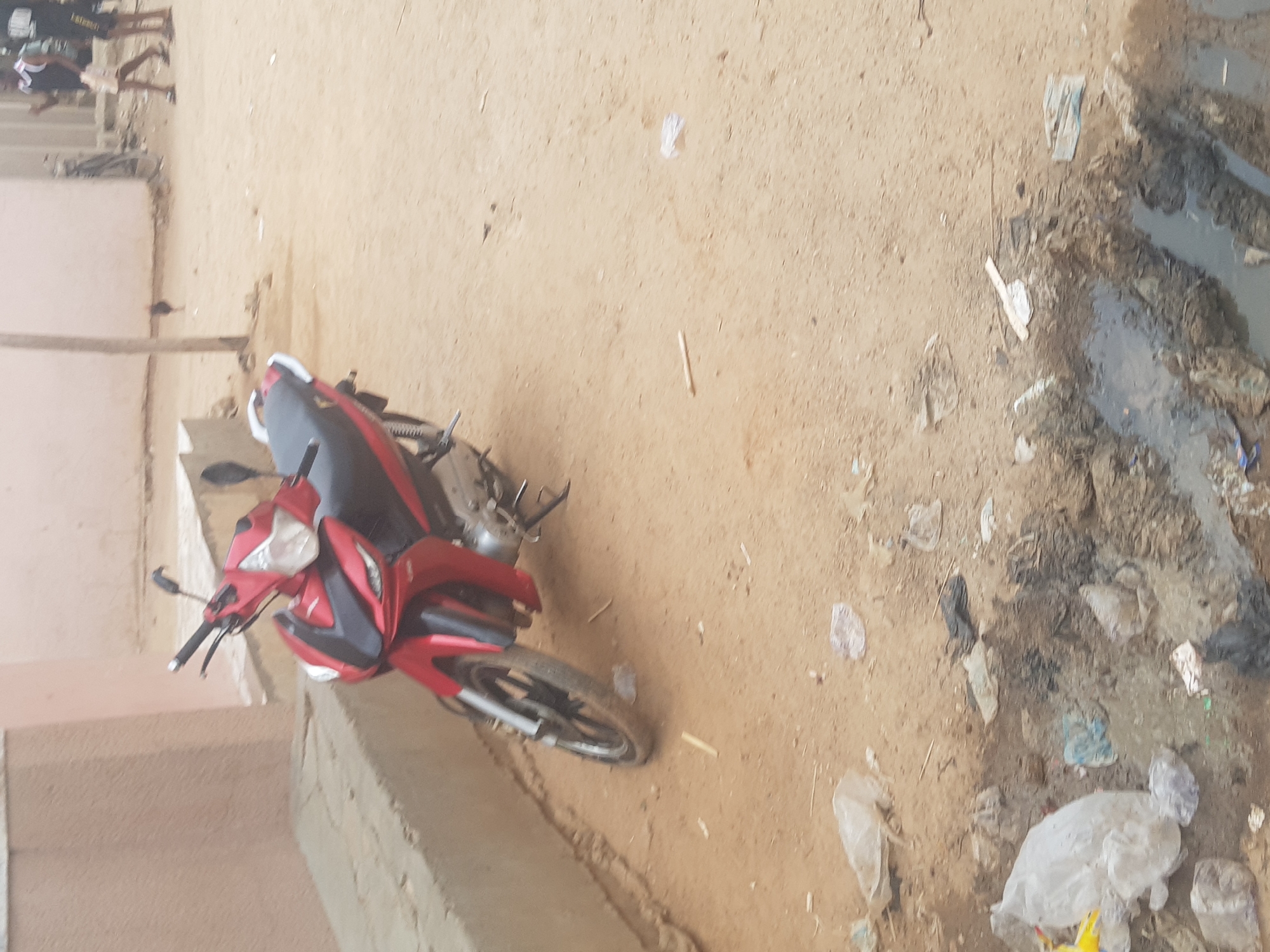 The image shows a red and black motorcycle parked on a dirt and concrete surface in Dutsinma B, Nigeria. The foreground includes trash and what appears to be a drainage ditch with stagnant water and debris. A partially visible building is on the left side of the image. The upper left corner shows a group of people in the distance.