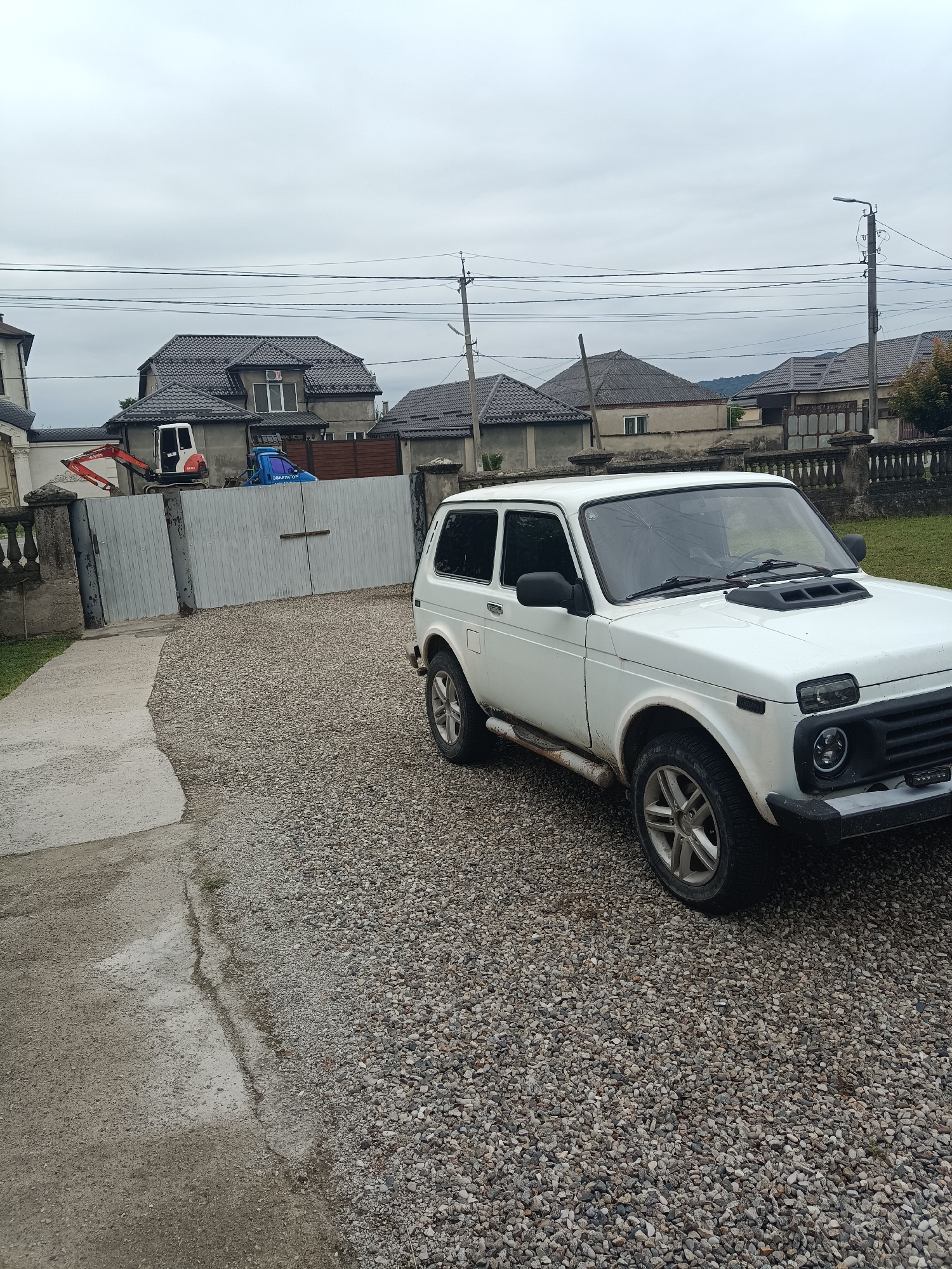 The image depicts a white Lada Niva parked on a gravel driveway in front of a closed metal gate. Behind the gate, a red excavator and a blue tow truck are visible. Several houses with tiled roofs are situated in the background under a cloudy sky. Utility poles and power lines traverse the sky. The environment suggests a residential area. The image appears to be taken in Kenzhe, Russia, as indicated by the context.