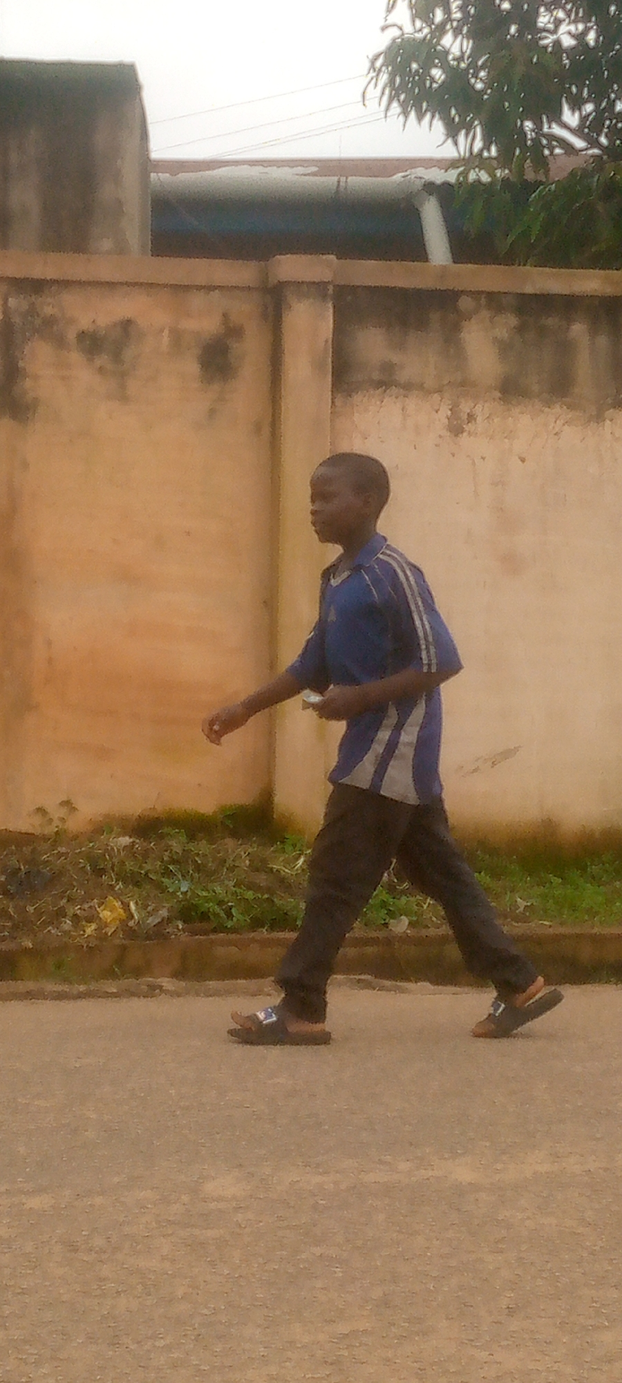A young boy walks on a paved road in front of a light brown wall. The boy is wearing a blue shirt with white stripes on the sleeves and dark pants. He has on dark sandals. The wall has some green vegetation growing at its base. In the background, the top of some buildings and trees are visible against a cloudy sky.