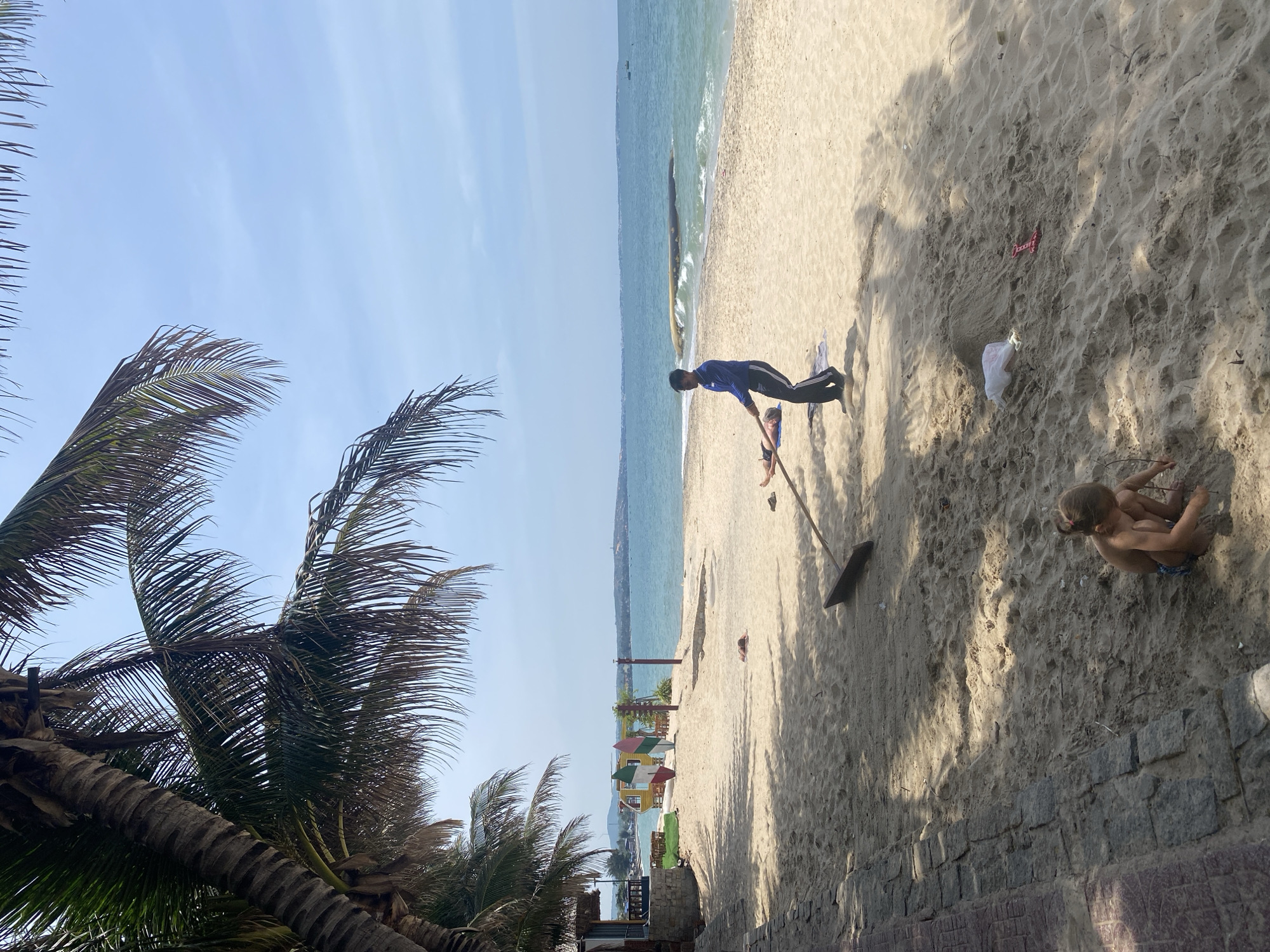 A sandy beach in Phu Thuy Ward, Vietnam, is depicted under a bright, overcast sky, with palm trees casting shadows across the foreground. In the middle ground, a man in a blue shirt and dark pants is carrying a long wooden pole, seemingly assisting another person lying on a towel in the sand. Further down the beach, a young girl is crouched in the sand, playing with a red plastic shovel. The ocean stretches to the horizon, with gentle waves lapping the shore. A few boats are visible in the distance. To the left, a building with a thatched roof and some colorful playground equipment suggest a resort or recreational area.