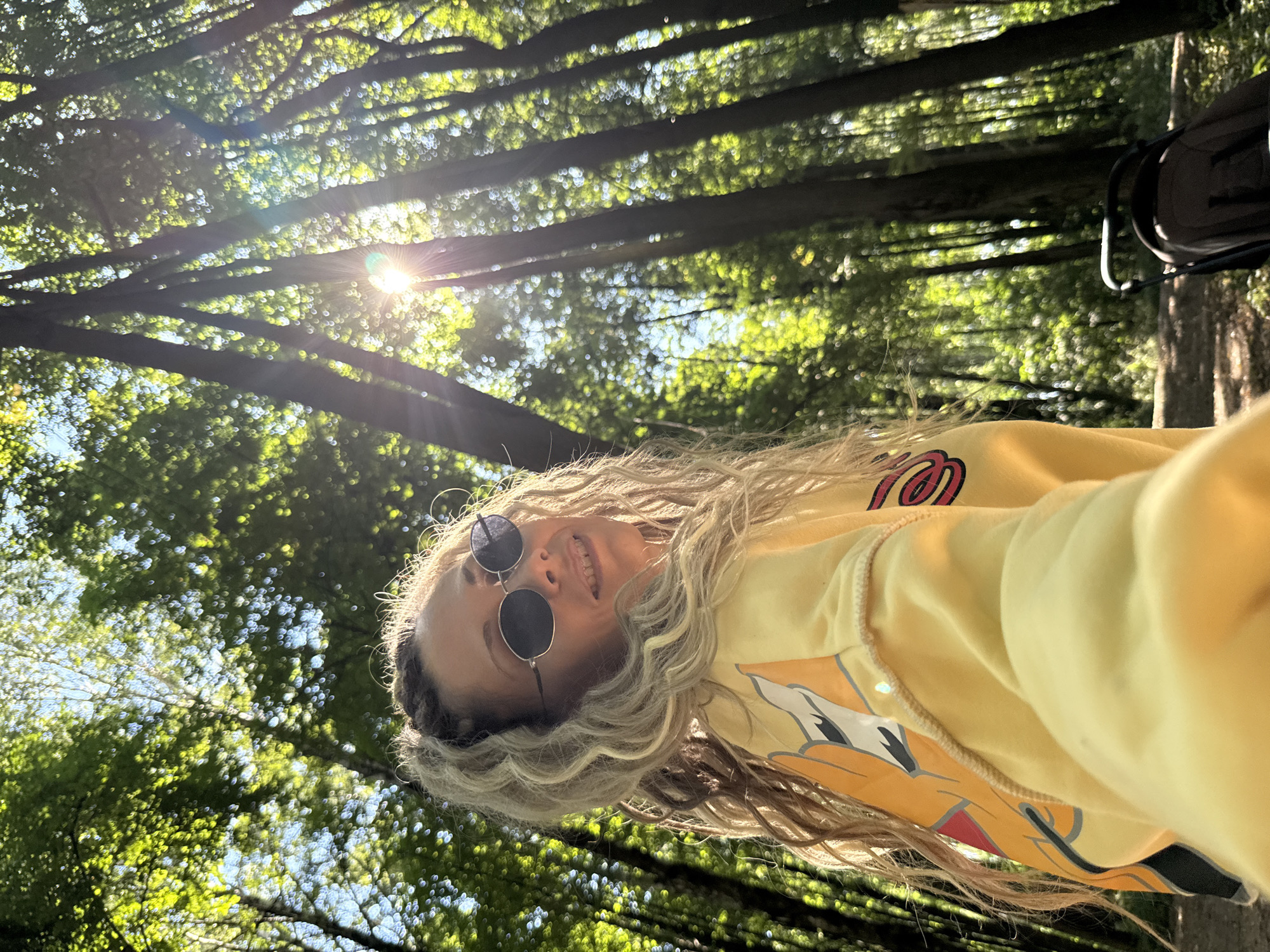 A woman with blonde, braided hair and sunglasses is taking a selfie in a sun-dappled forest. She is wearing a yellow sweatshirt with a graphic print on the sleeve. The background shows tall trees with sunlight filtering through the leaves. A stroller is visible in the lower right corner. The location is the city of Rusanovo, Russia.