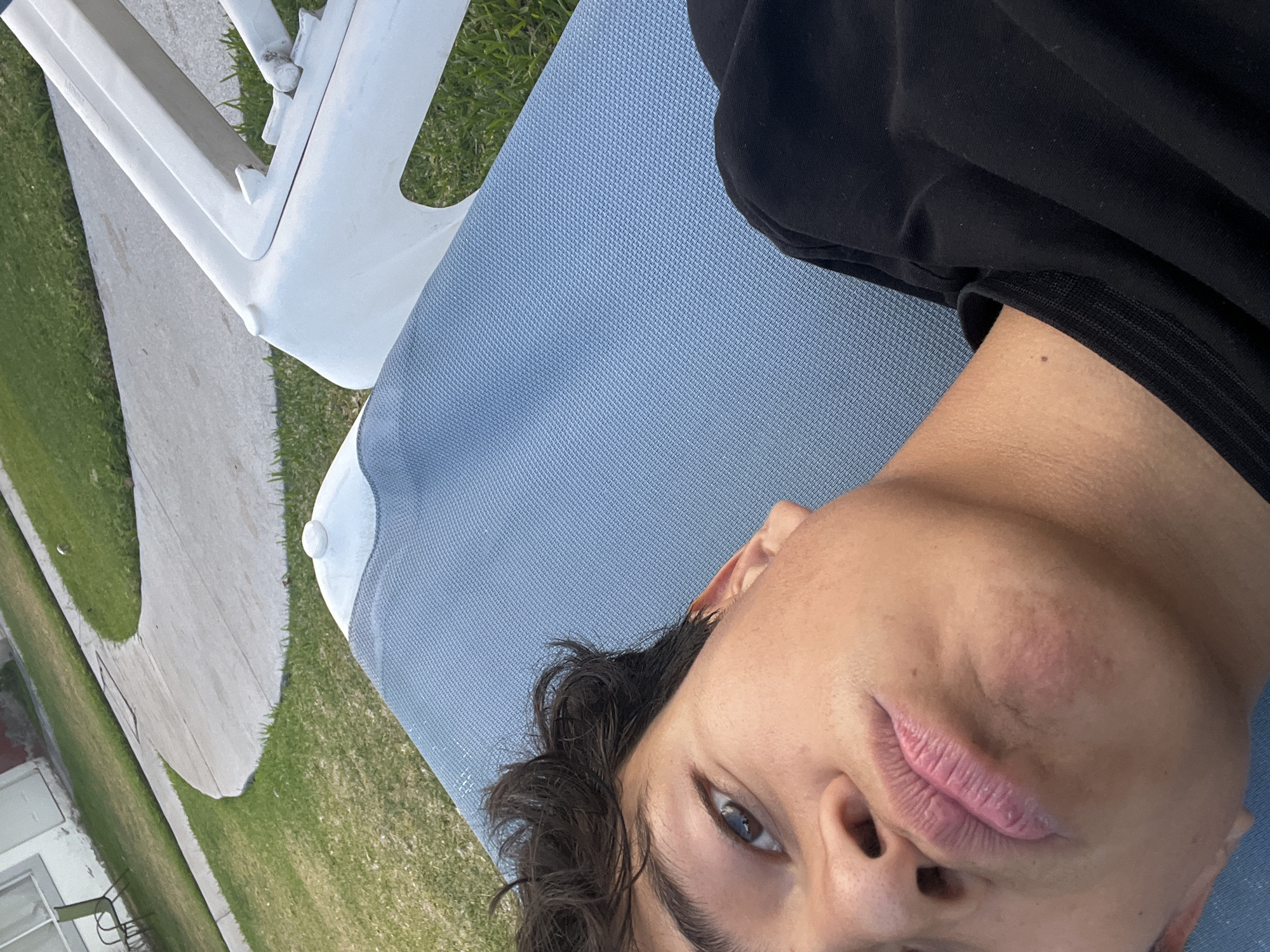 A person lies on a blue-mesh chaise lounge outdoors, partially visible. The person is wearing a black long-sleeved shirt. Only a portion of the person's face is visible. The chaise lounge is positioned on grass, adjacent to a concrete sidewalk. A white plastic chair or table leg is also present. The location is inferred to be Xochitepec, Mexico.