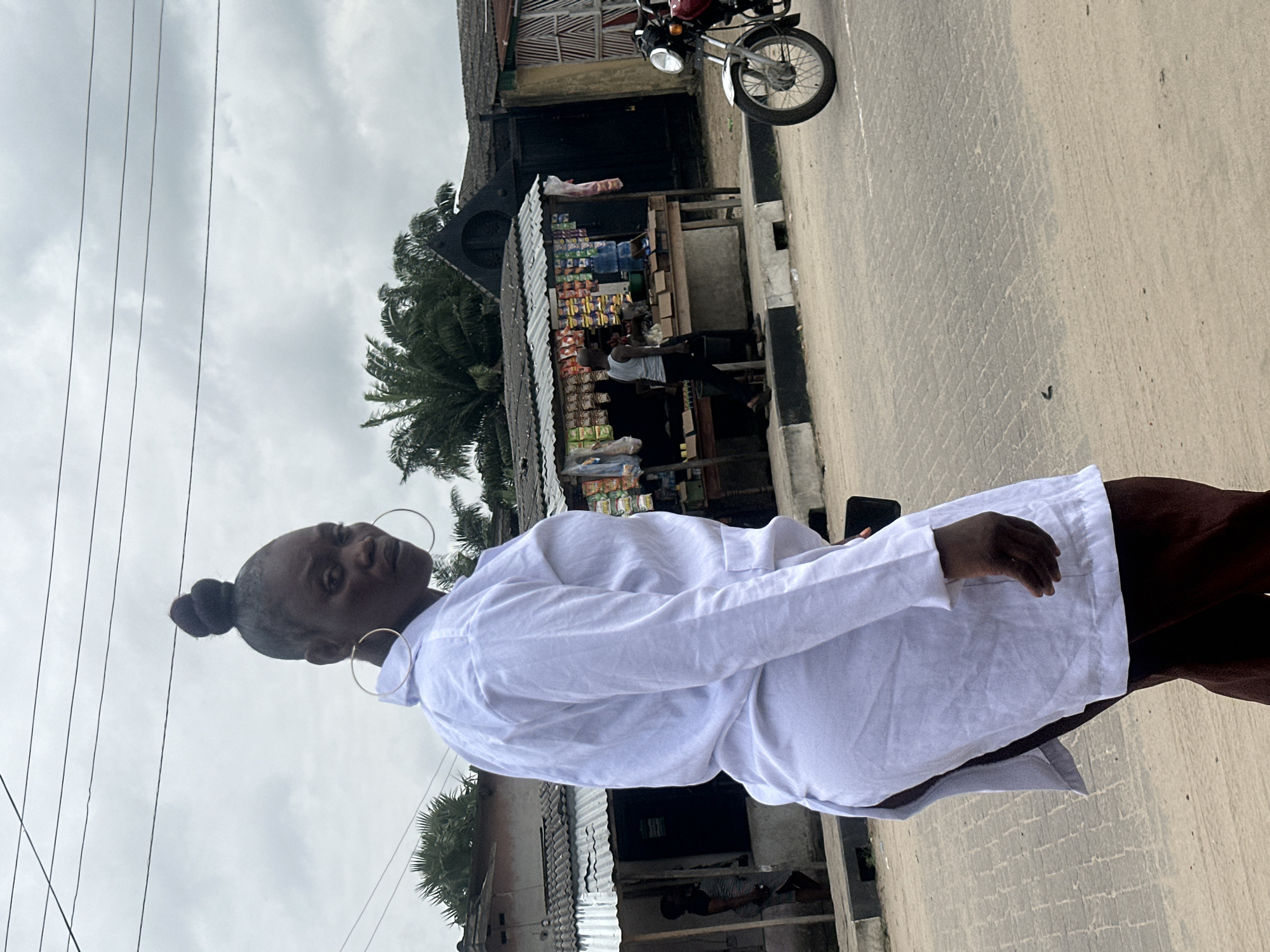 A woman wearing a white lab coat and dark pants walks on a paved street in Badagry, Nigeria. She has her hair pulled up in a bun and is wearing large hoop earrings. She is looking back over her shoulder at the camera. In the background, there are small shops with corrugated metal roofs and a motorcycle. The sky is overcast.