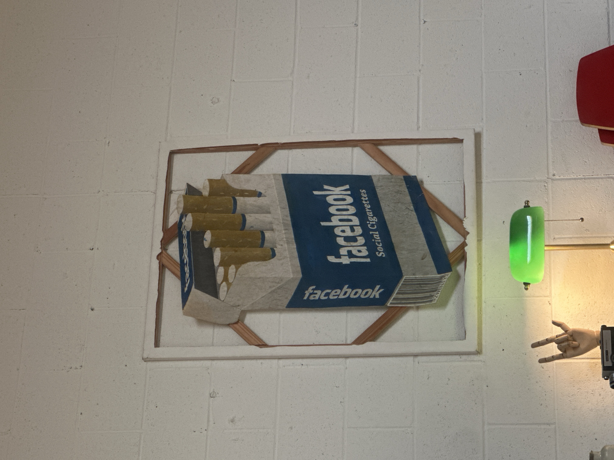 The image shows a framed piece of art hanging on a white brick wall. The artwork depicts a pack of cigarettes with the "facebook" logo and the words "Social Cigarettes" printed on it. The "cigarettes" themselves are visible at the open end of the pack. To the right of the artwork, there is a red lamp fixture and a green lamp. Also visible is a wooden hand posed in a "rock on" gesture. The image does not provide sufficient information to determine the location of the art in the city of Glendale, United States of America.