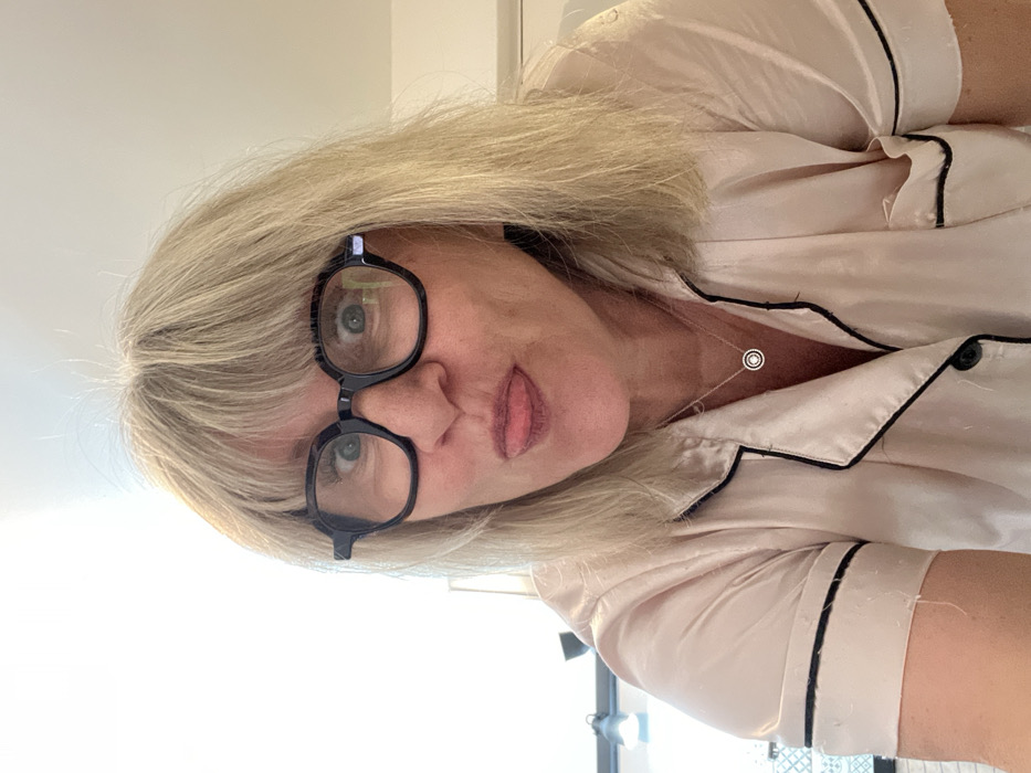 A woman with blonde hair and glasses looks directly at the camera. She wears a light pink pajama-style top with black piping. A delicate necklace with a small pendant is visible. The background is mostly white, hinting at a bright interior space. Part of a lighting setup can be seen in the background, suggesting the photo may have been taken indoors with studio lights.