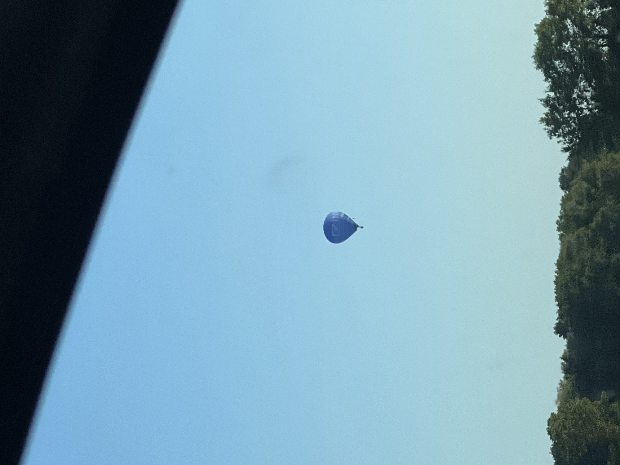 A single blue hot air balloon floats in a clear blue sky above a dark green treeline. The balloon appears to be advertising or carrying a message, with text visible on its surface, although it is not clearly legible. A dark, angled shape, likely a car roof or a similar obstruction, partially obscures the top of the image.