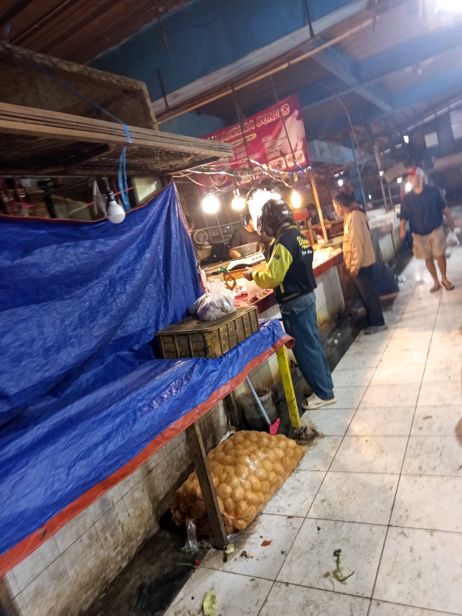 The image depicts an outdoor market scene, likely in Karawang, Indonesia, given the context. The foreground shows a large blue tarp covering a stall or structure, with a red trim along the bottom edge. To the right of the tarp, a large mesh sack filled with potatoes is visible, resting on the tiled floor. The floor itself appears somewhat grimy and wet in places.
In the midground, several individuals are present. A person wearing a yellow and black jacket and a helmet is standing behind a counter, appearing to interact with produce or goods. Another person in a tan jacket is standing to their right, looking towards the counter. Further to the right, a man in a dark blue shirt and light-colored shorts is walking away from the viewer. The market is covered by a wooden structure with exposed beams and a corrugated metal roof. Several light bulbs are illuminated, suggesting it is evening or dimly lit. A banner with text in Indonesian hangs in the background. The overall impression is a busy, somewhat rustic market environment.