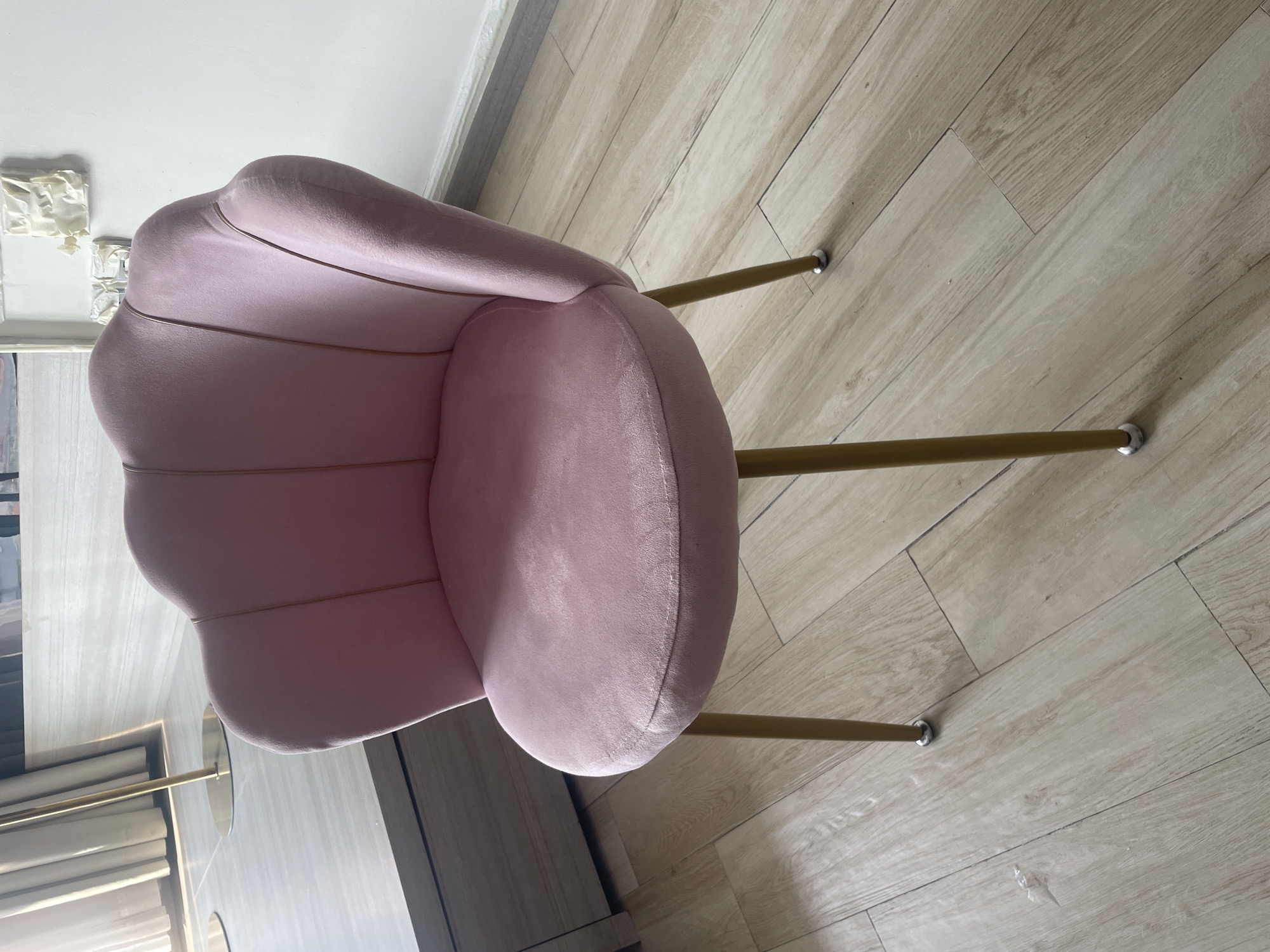 A high-angle shot shows a pink velvet chair with gold-colored metal legs on a light wood-look tiled floor. The chair has a scalloped back and a round seat. The background includes part of a white wall and some furniture. The location appears to be indoors. The scene is not clearly indicative of being in Bắc Nha Trang, Vietnam, so information to the contrary is needed.