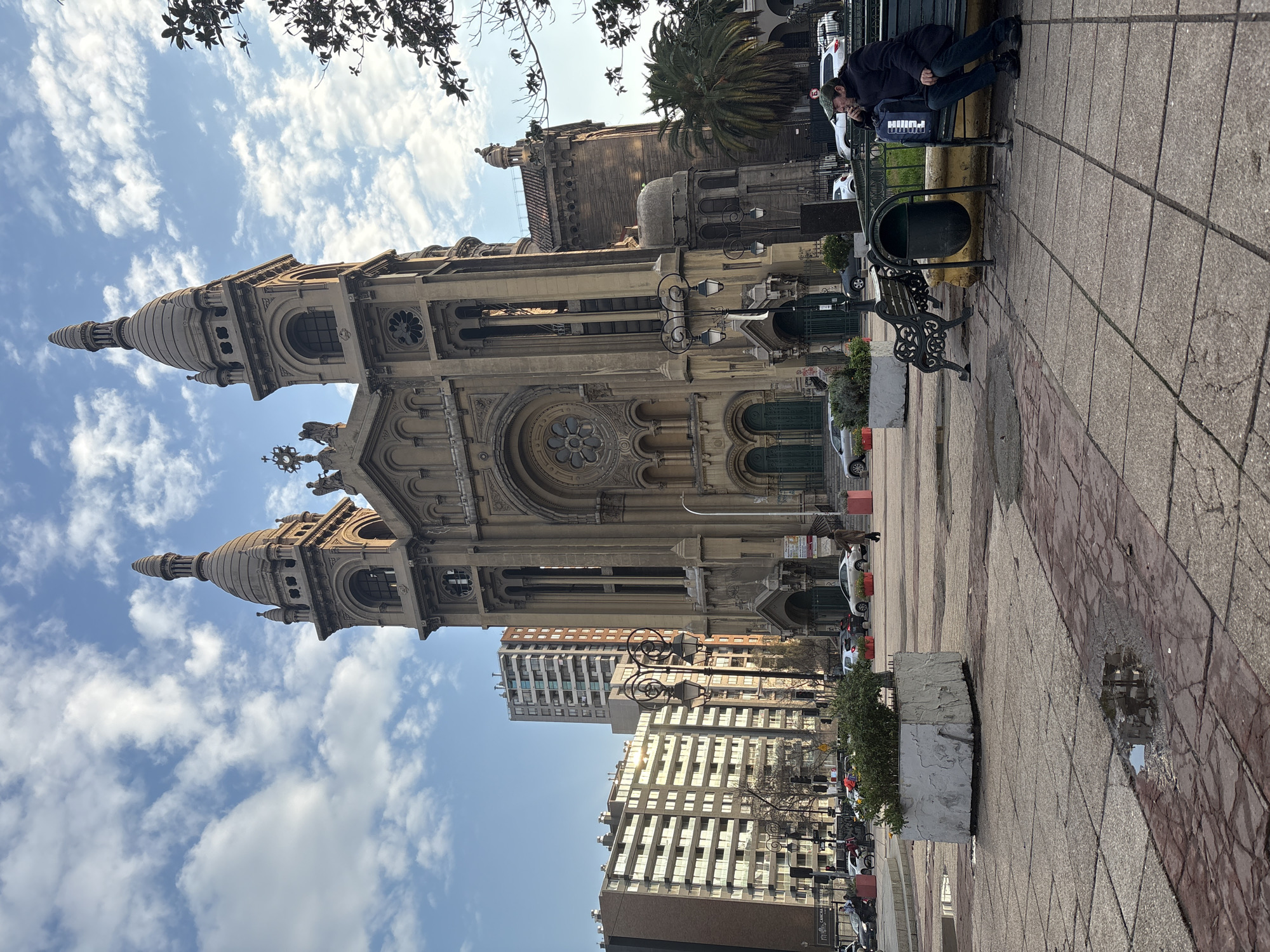 The image captures a view of the Metropolitan Cathedral of Santiago, Chile, on a cloudy day. In the foreground, a man is seated on a park bench, looking to his right and holding a cigarette. He is wearing a dark jacket and jeans. A dark blue backpack with white lettering is placed next to him on the bench. To his left is a black trash can. The bench is situated on a paved plaza with a pattern of square tiles. In the mid-ground, a large planter box with green foliage is visible, and beyond that, a street with cars and pedestrians. Behind the plaza and the street are several modern, multi-story buildings. The Metropolitan Cathedral is a grand, historic building with two prominent towers and an ornate facade. The sky is blue with scattered white clouds.