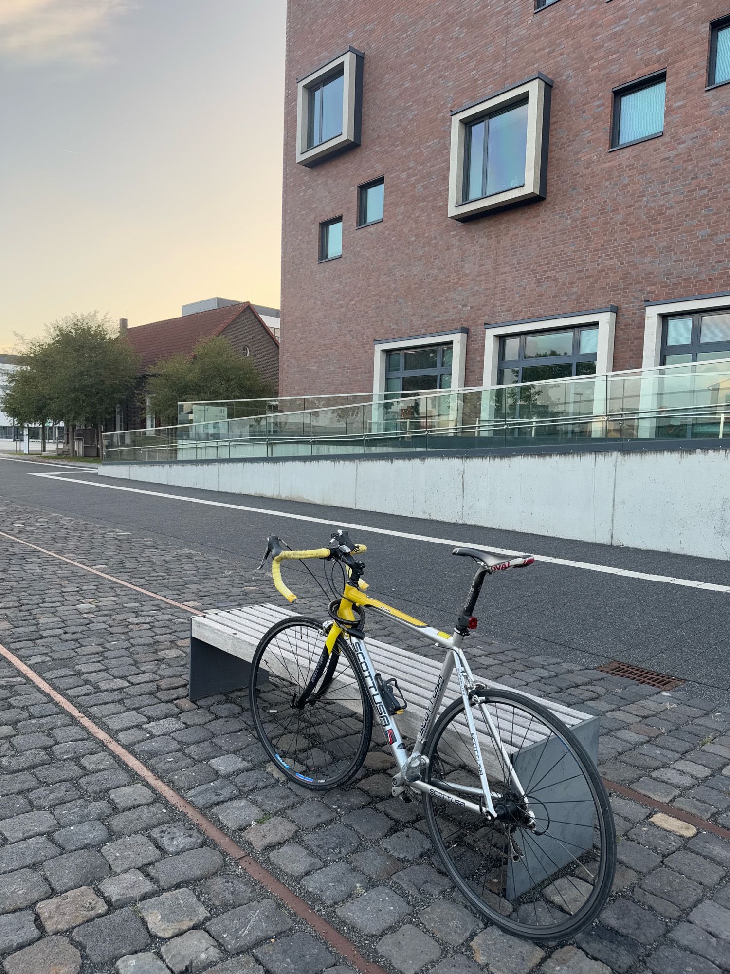 A silver and yellow road bicycle is parked on a cobblestone path in front of a modern brick building with large windows. The bicycle is leaning against a wooden and concrete bench. The building appears to be an office or residential building, with a clean, contemporary design. The sky is overcast, and the overall lighting suggests it is late afternoon or early evening. In the background, there are trees and another smaller building with a red tiled roof. The scene is devoid of any people. The location appears to be an urban or semi-urban area.