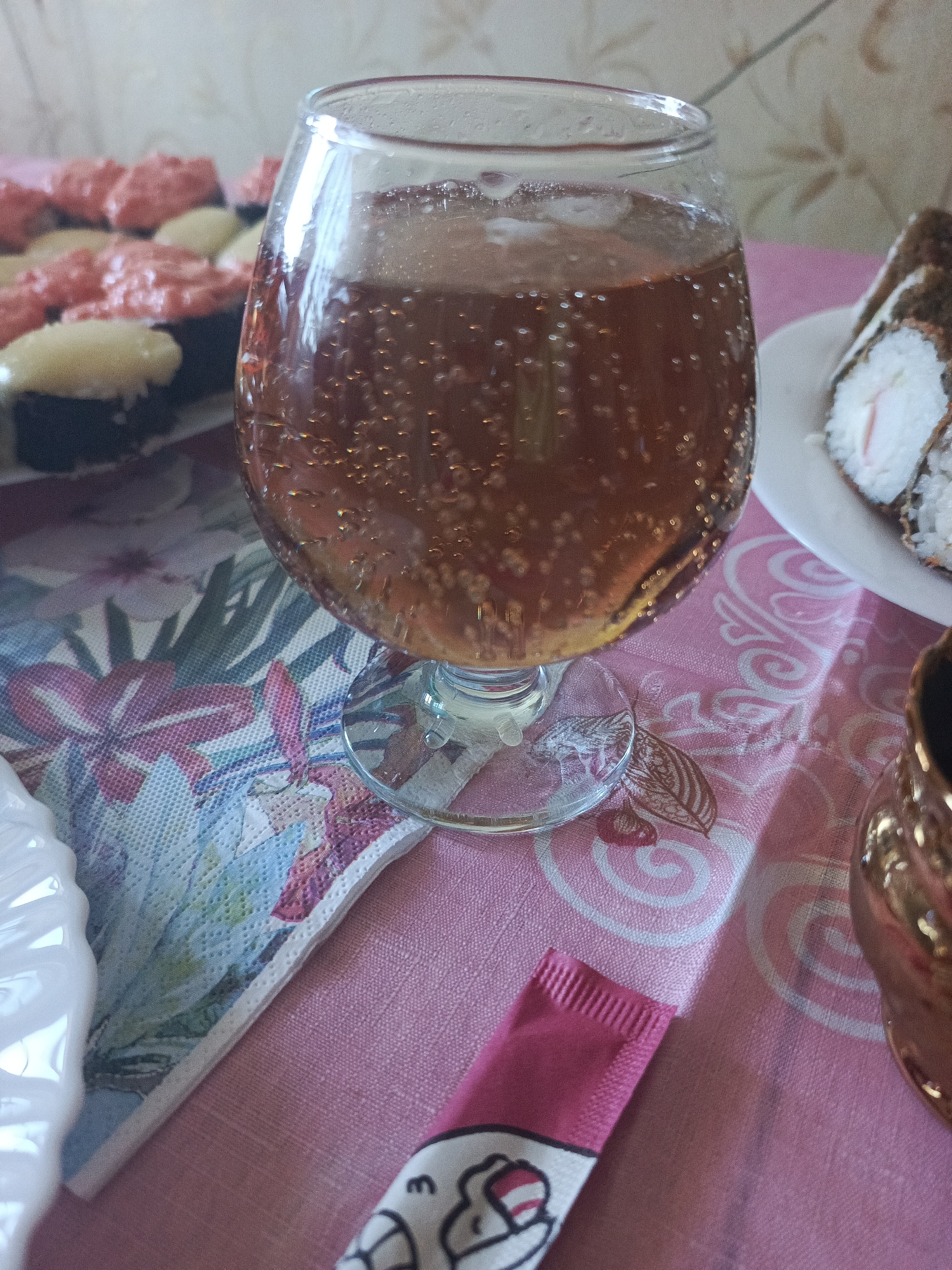 A close-up, eye-level shot shows a glass of a bubbly, amber-colored beverage on a pink tablecloth. The glass is partially filled and has many small bubbles rising. To the left and slightly behind the glass are several pieces of sushi, topped with pink sauce and white sauce. To the right of the glass, a white plate holds a single piece of sushi roll. In the foreground, a pink packet with black cartoon drawings is visible. The background is out of focus, with a wall exhibiting a pattern of beige and brown shapes. The overall scene appears to be a casual meal setting.