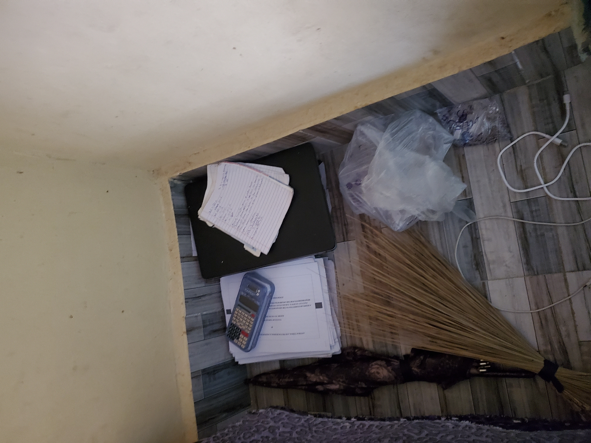 The image depicts a corner of a room with tiled flooring and plain walls. On the floor, a stack of papers is topped with a calculator. Adjacent to the papers is a closed laptop. To the left of the papers, a dark, patterned object, possibly an umbrella or a rolled-up textile, lies on the floor. A broom with a dark handle and a straw head is positioned in front of the umbrella and extends towards the foreground. In the center of the image, a clear plastic bag filled with white material is visible, with another smaller bag containing small, dark objects next to it. A white cable lies on the floor in the lower right quadrant of the image. The overall scene suggests a cluttered storage area or a workspace. The image is not specific enough to determine its exact location within Jalingo, Nigeria, but it could be a room in a residential or commercial building.