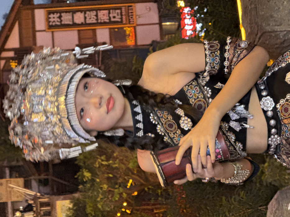 An East Asian female is depicted in traditional attire, standing outdoors at night in the Hongyadong complex, Yuzhong District, Chongqing, China. She wears an elaborate silver headdress composed of numerous metallic pieces and dangling elements, silver dangle earrings, and a black two-piece garment adorned with intricate silver embroidery, colorful patterns, and metallic embellishments. Glitter is applied to her cheeks, and her dark hair is styled in two long braids. On her left wrist, she wears a silver cuff bracelet with dangling chains. She holds a small, reddish-brown cylindrical hand drum with both hands. Her gaze is directed towards the viewer with a slight smile. The background features blurred traditional multi-story Chinese buildings, illuminated by artificial lights, with a prominent vertical sign reading "洪崖洞酒店" (Hongyadong Hotel) and "HOTEL". Green foliage with lights is visible to the left, and additional red and white lights are blurred in the distance.