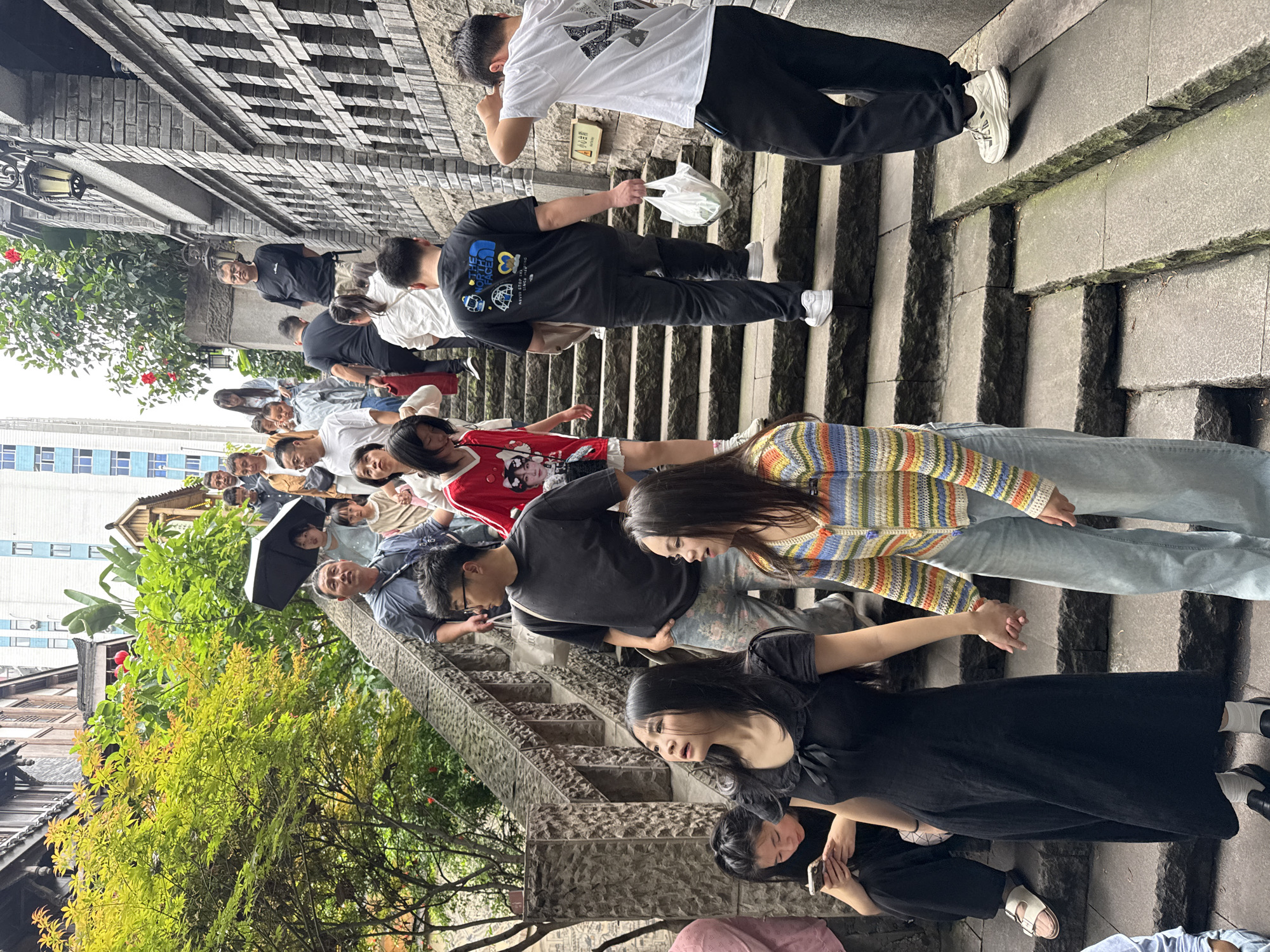 A daytime scene shows a crowd of people walking up a gray stone staircase in the city of Yuzhong District, China. The staircase has a rough-hewn texture and is flanked by stone walls and greenery. The crowd is diverse in age and attire, with some people holding umbrellas. Trees and building architecture are visible, blending nature with urban elements. Some individuals look at their phones.